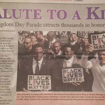 People marching in a Black Lives Matter parade, holding signs that say 'BLACK LIVES MATTER' and wearing business suits. The parade is in a city street with a crowd in the background.