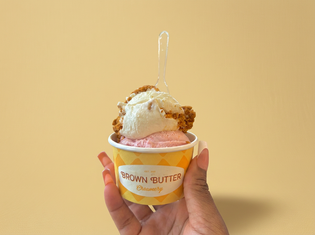 Hand holding a cup of ice cream with two flavors, topped with a caramel drizzle and cookie pieces, against a yellow background.