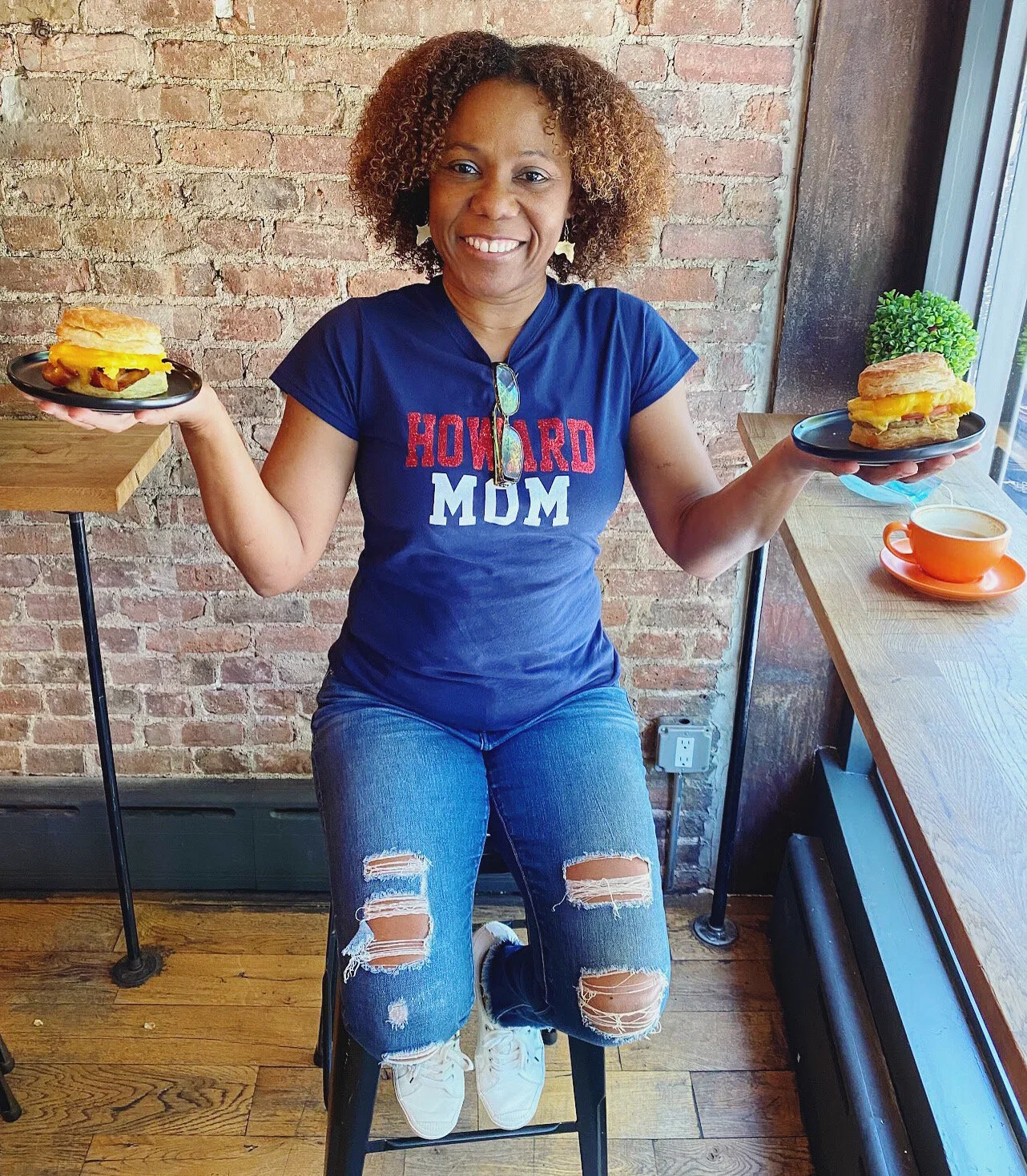 A woman with curly hair wearing a navy blue T-shirt that says 'Howard Mom' and ripped jeans, sitting on a black chair in a cafe, holding plates with breakfast sandwiches, with a brick wall behind her and a cup of coffee on the table next to her.