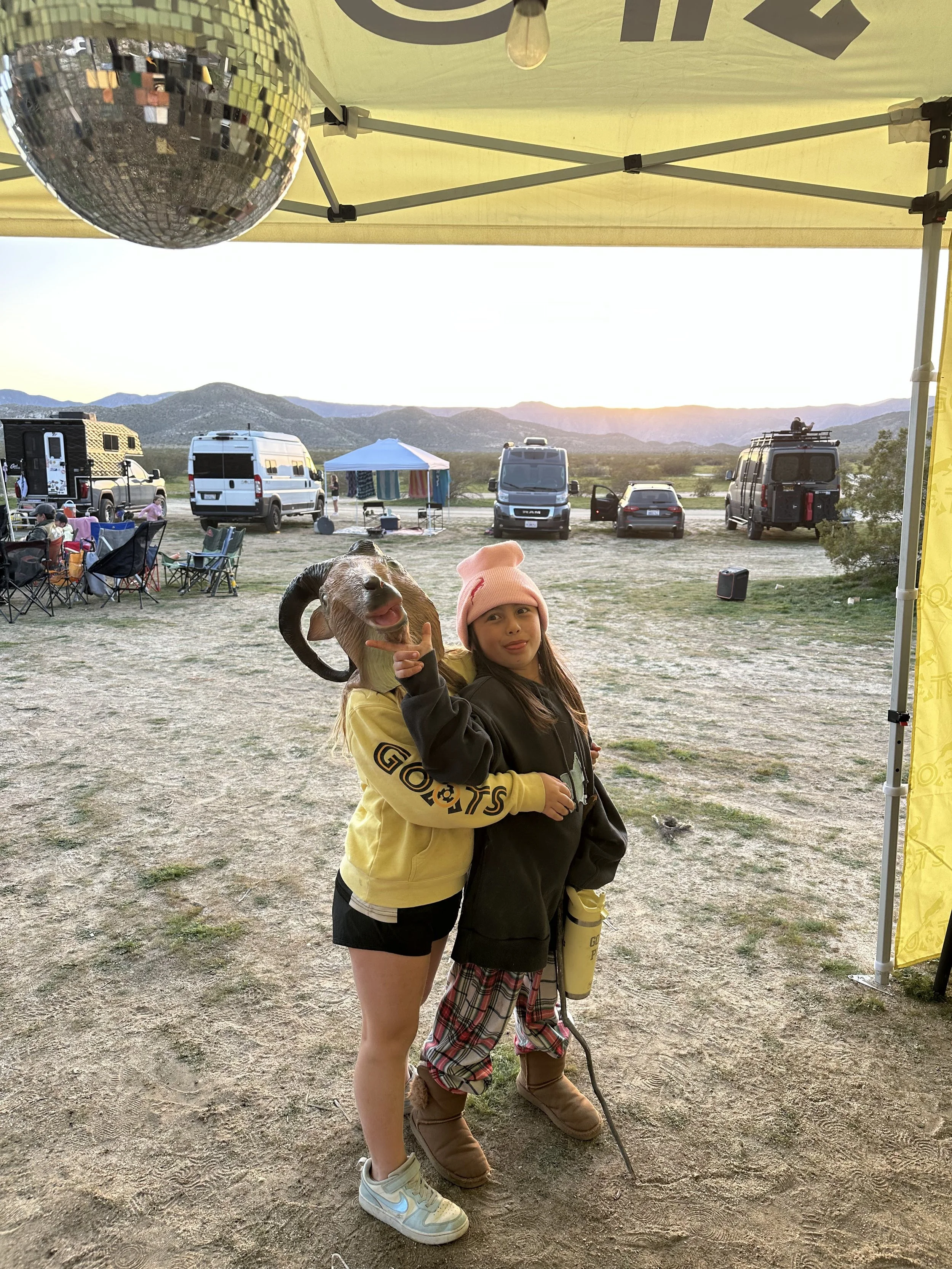 Two young girls standing under a yellow canopy tent outdoors at sunset, holding a goat with one girl pointing at it. The girl on the left is wearing a yellow hoodie, shorts, and sneakers. The girl on the right is wearing a pink beanie, plaid pants, and brown boots. In the background, there are camper vans, a small tent, chairs, and mountains.