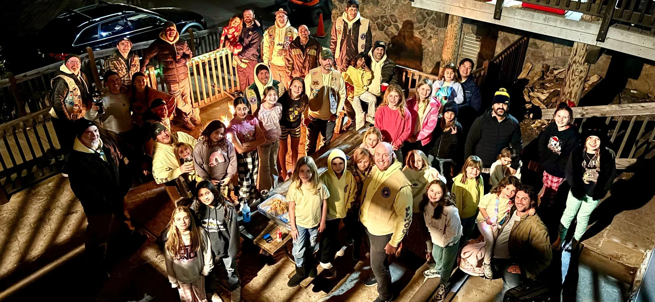 A large group of people, including children and adults, gathered outdoors at night on a decking area, posing for a group photo. There is a table with snacks, and some individuals are holding drinks. The scene is illuminated by nearby lights, and a stone wall and parked cars are visible in the background.
