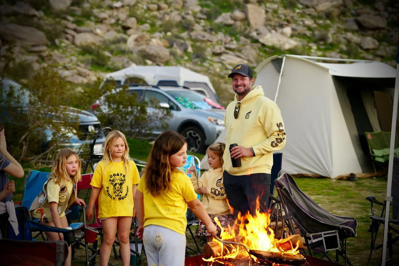 A group of kids wearing yellow 'Goats Family Reunion' shirts gathered around a campfire with an adult man in a yellow hoodie, all outdoors with tents, cars, and rocky hills in the background.