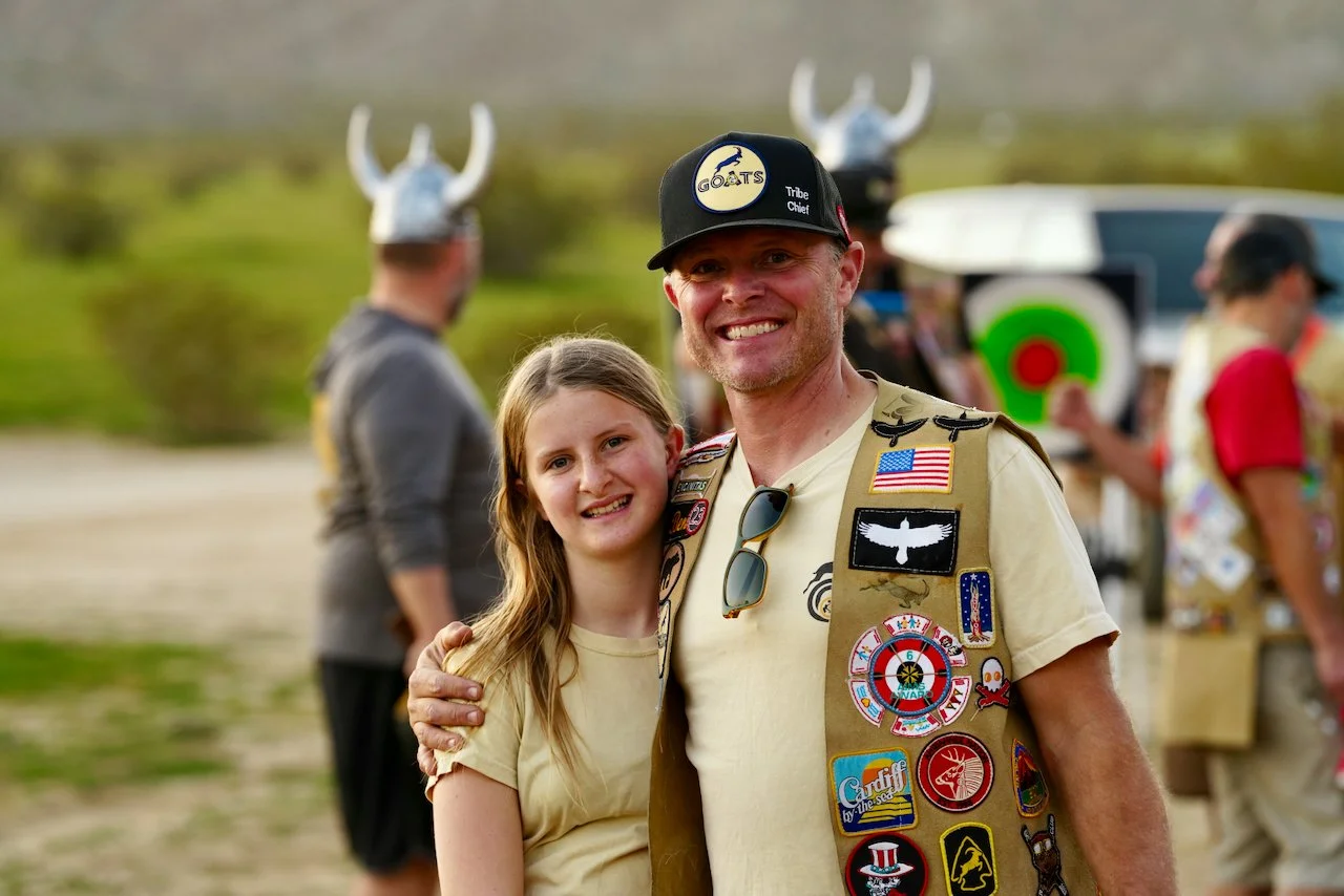 A man in a military vest decorated with patches, smiling and standing next to a young girl, during an outdoor event with other people in the background, some wearing Viking helmets.