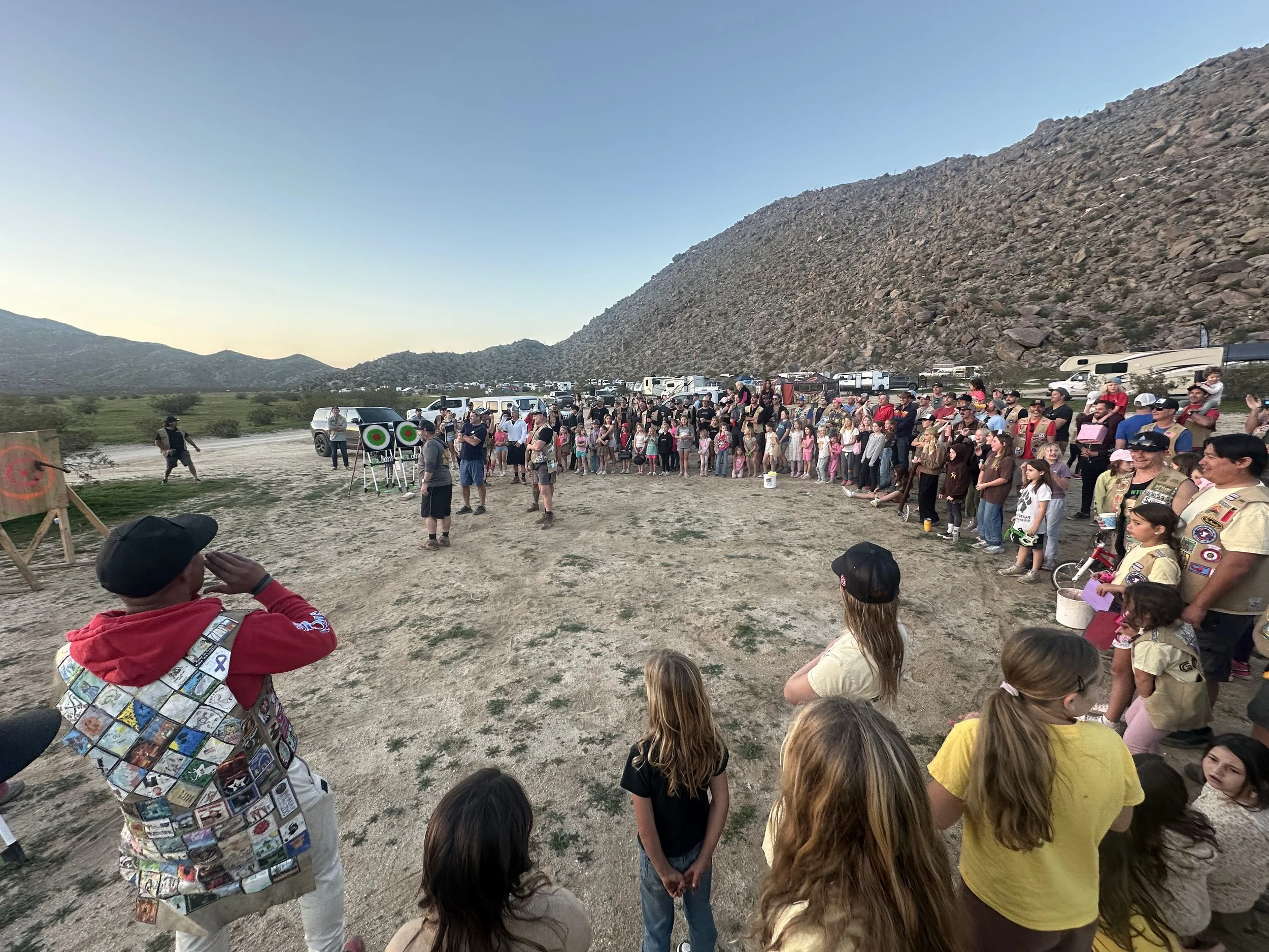 A large group of people gathered outdoors at a shooting range, aiming at targets, with a mountainous landscape in the background at sunset.