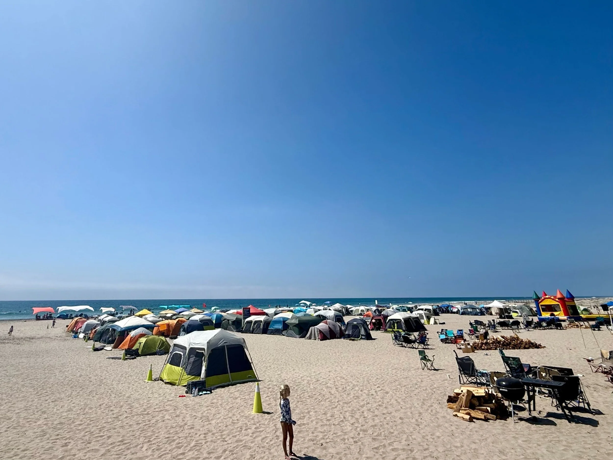 A crowded beach with numerous tents and umbrellas set up on the sand, under a blue sky, with some people walking and playing, and a few structures and chairs visible in the distance.
