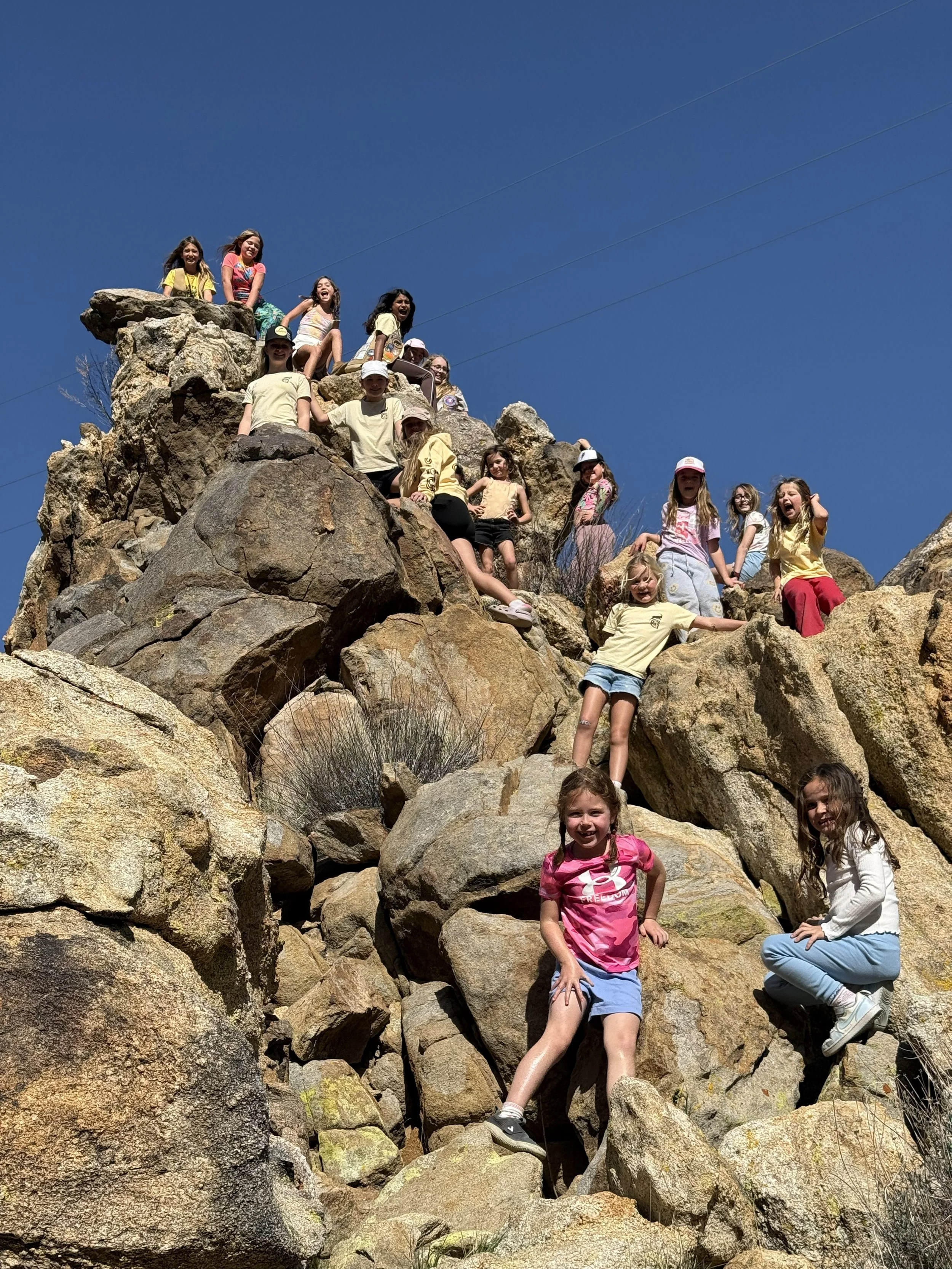 Group of children climbing and playing on large rocks outdoors under a clear blue sky.