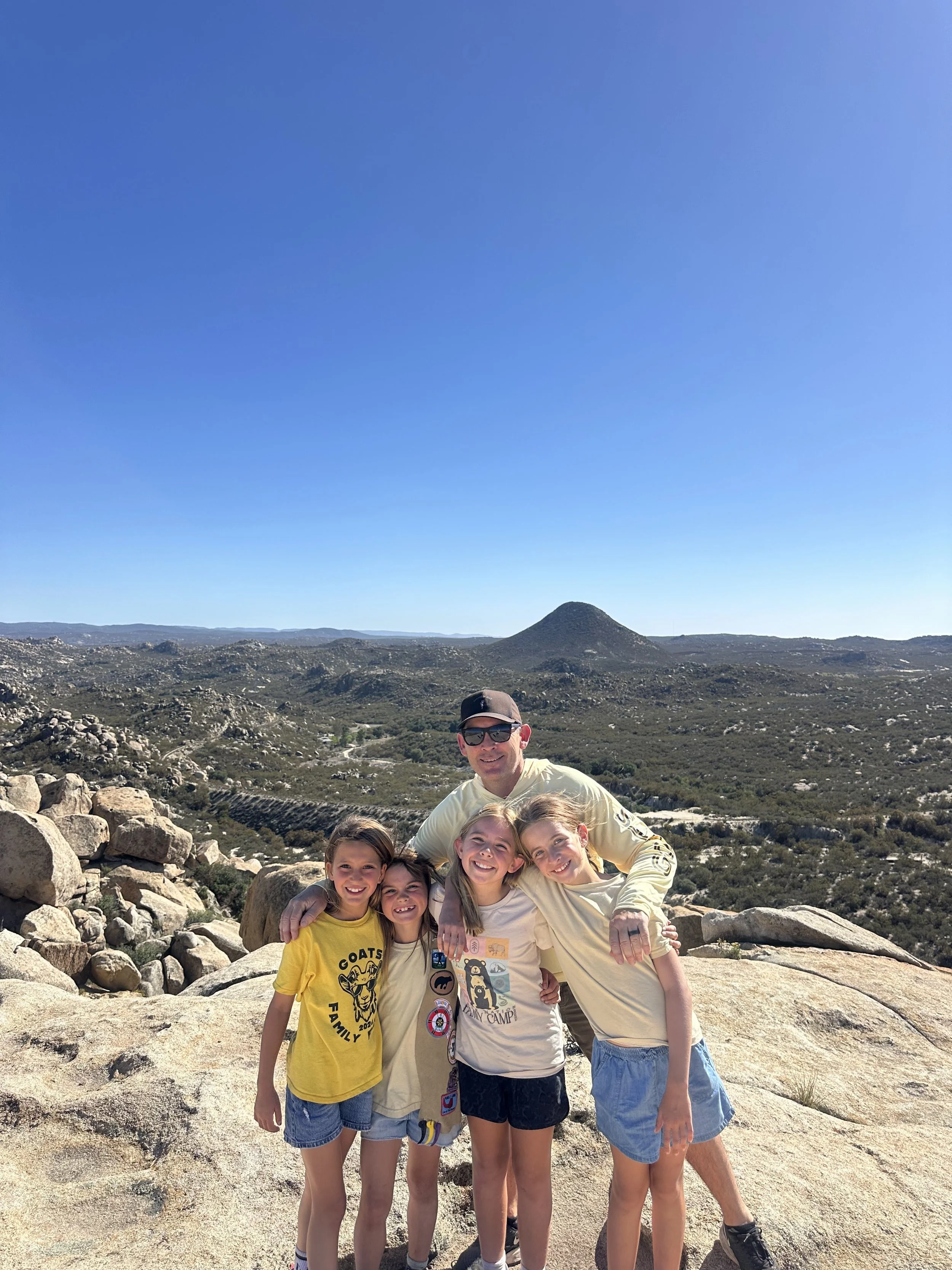 A group of five people—three young girls, a teenage girl, and an adult man—are smiling and posing for a photo in a rocky desert landscape with a mountain in the background under a clear blue sky.