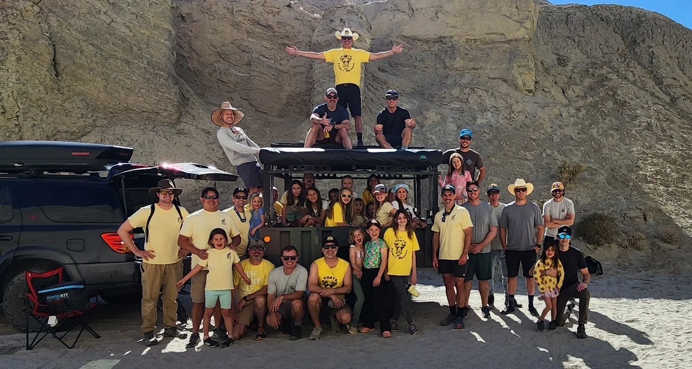 Group of people posing in front of a rocky desert landscape, some standing on a vehicle and some sitting or kneeling on the ground, with clear blue sky above.