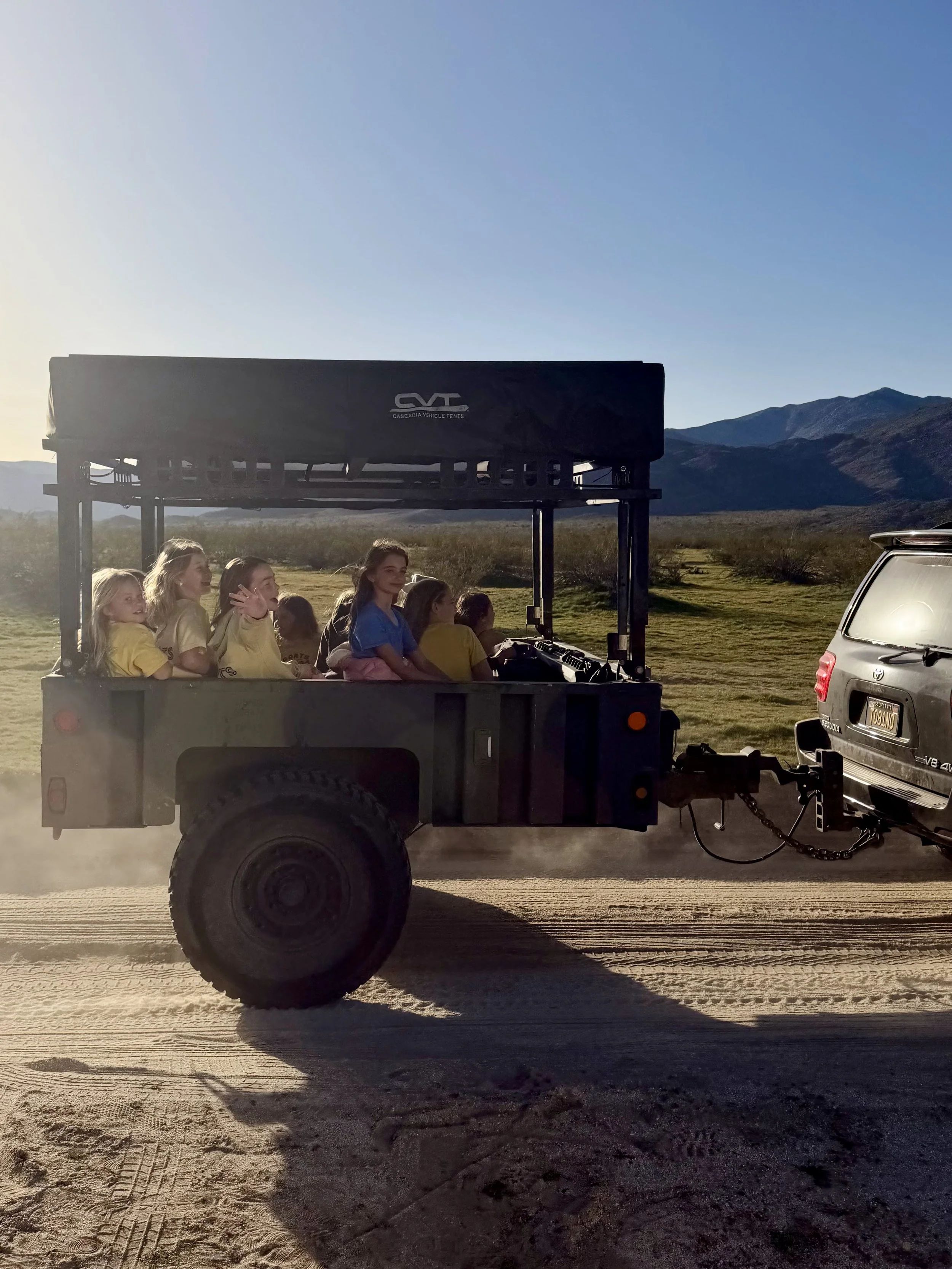 Group of people riding in a utility vehicle with a canopy, traveling on a dirt road in a rural landscape with mountains in the background during sunset.
