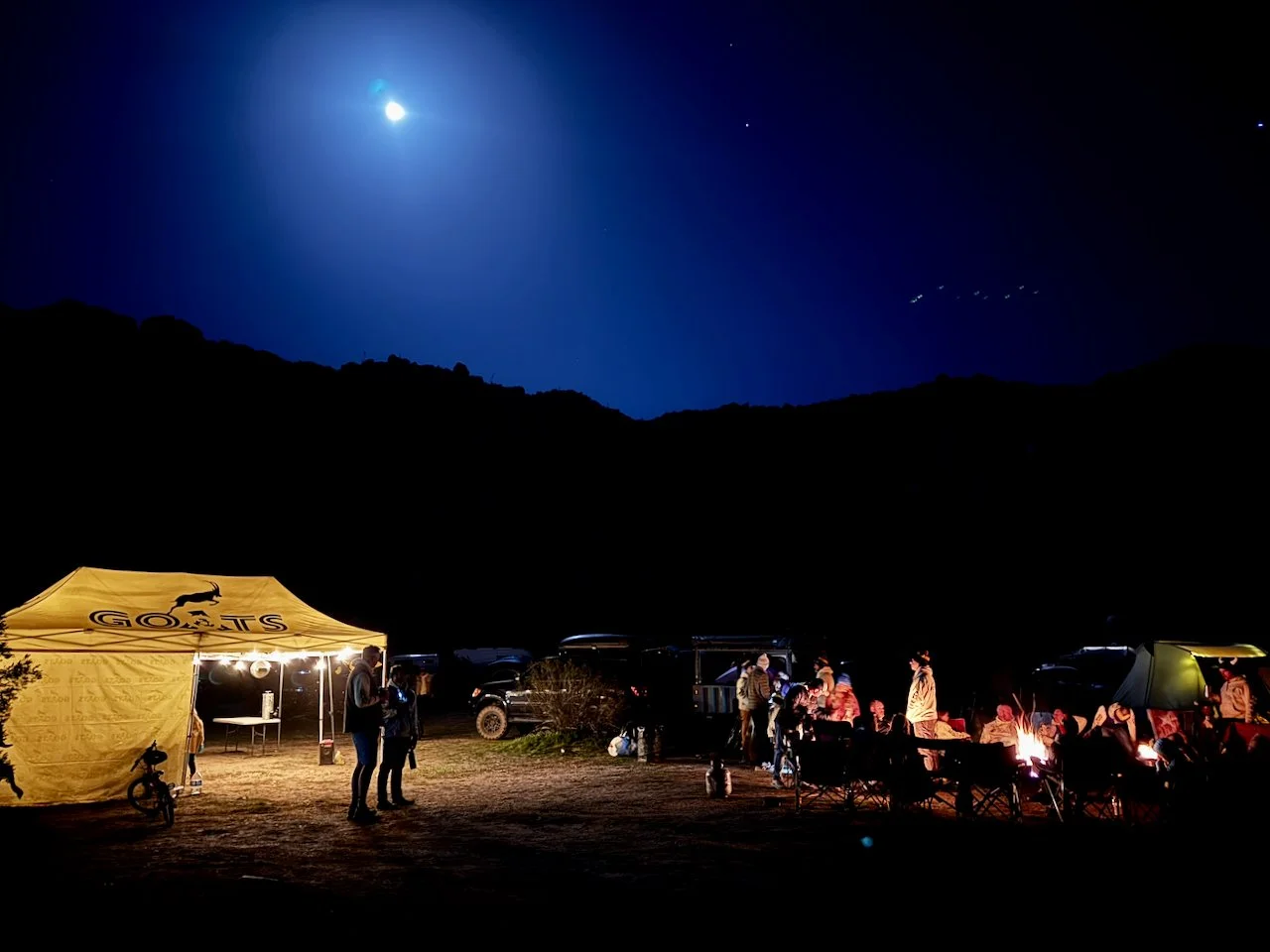 Nighttime outdoor gathering with a group of people around a campfire, tents, and vehicles under a dark sky with a bright moon and stars.