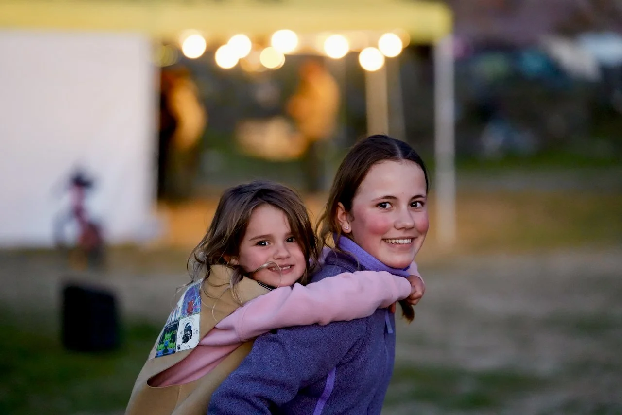 Two smiling girls, one giving the other a piggyback ride, outdoors during sunset.