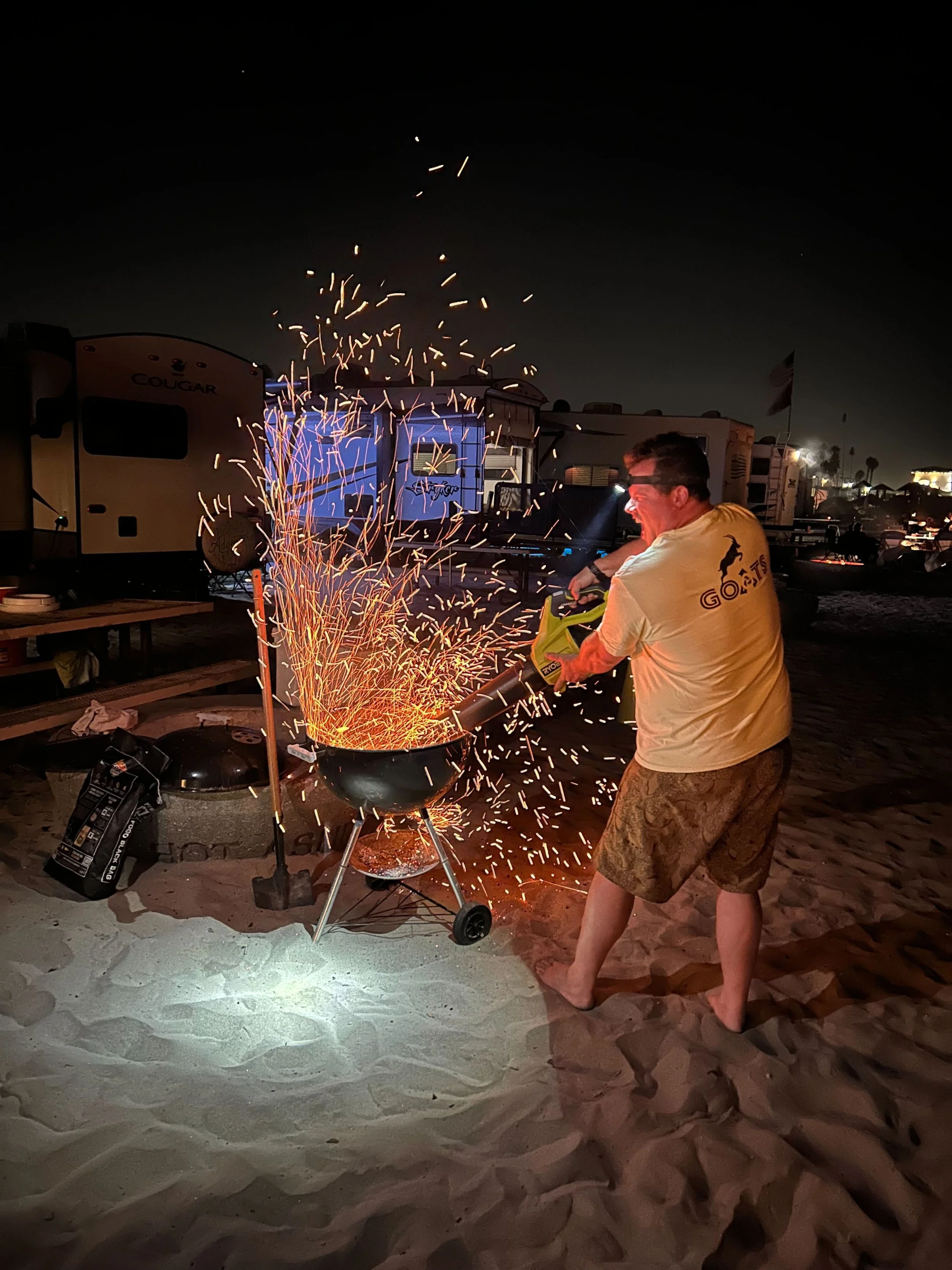 A man in a yellow t-shirt and patterned shorts using a fireworks device on a barbecue grill at night on a sandy beach, with sparks flying, RVs, tents, and flags visible in the background.