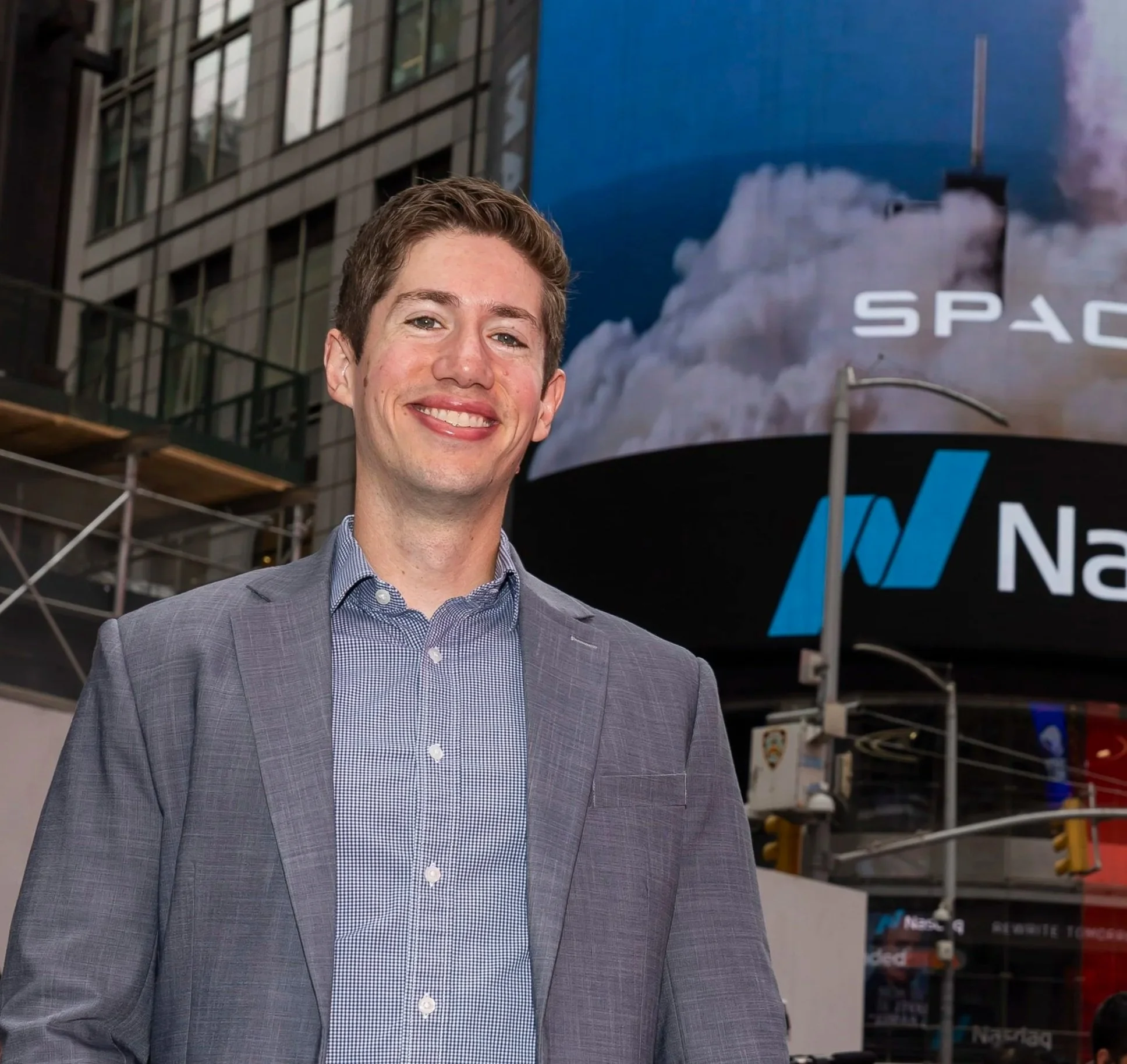 Smiling young man in a gray suit jacket and checkered shirt standing outdoors in front of a large digital billboard at night, with city buildings in the background.