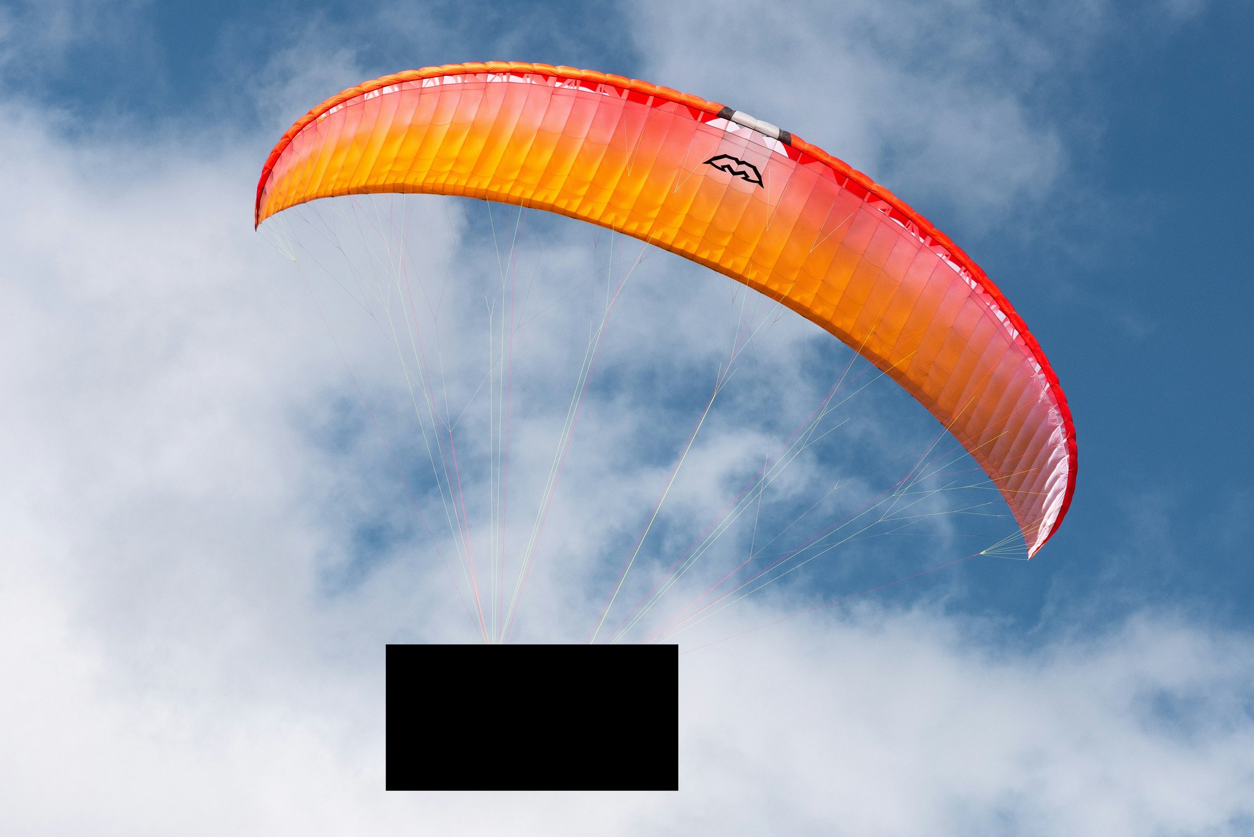 A colorful orange and yellow kite flying in a cloudy blue sky. A black box obscures a prototype drone design.