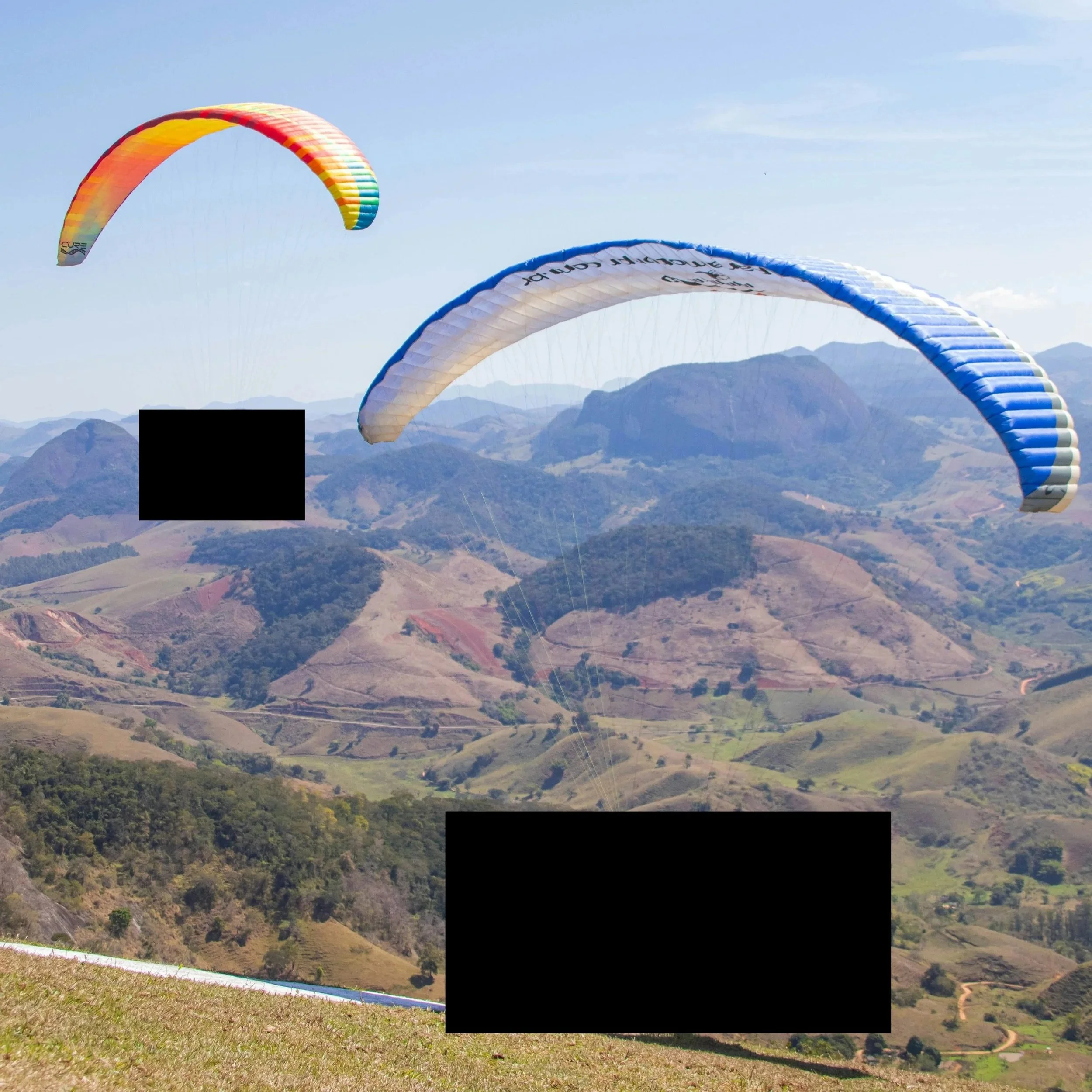 Two colorful paragliders soaring over a mountain landscape with hills and valleys, under a clear sky. Black boxes obscure prototype drone designs.