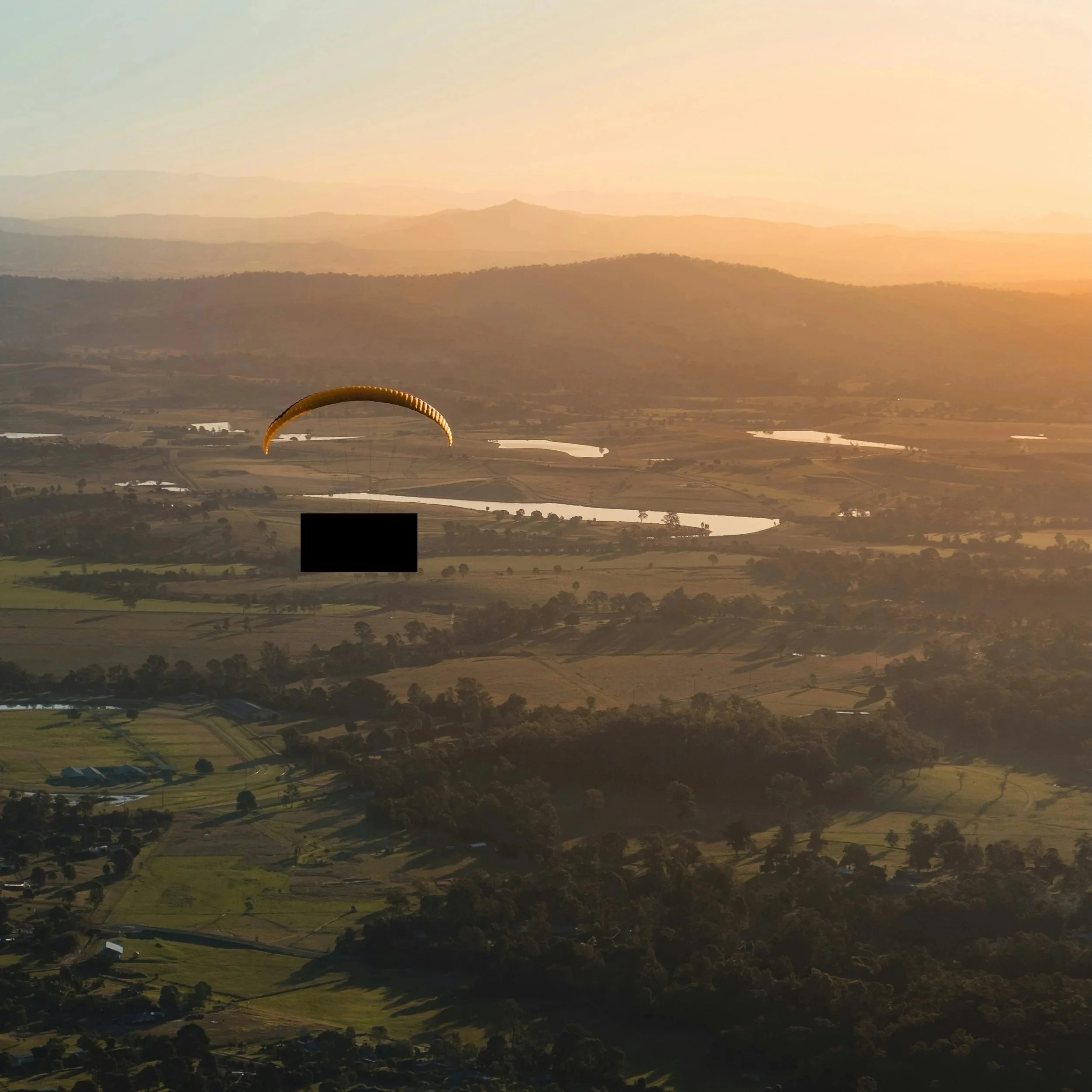 A landscape view of rolling hills and farmland at sunset, with a paraglider wing visible in the lower part of the image. A black box obscures a prototype drone design.