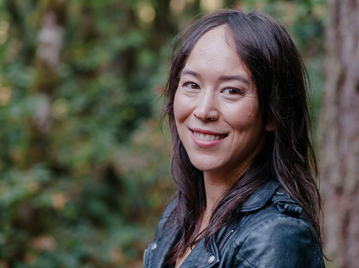 A woman with dark hair and a nose ring smiling outdoors with a blurred forest background.