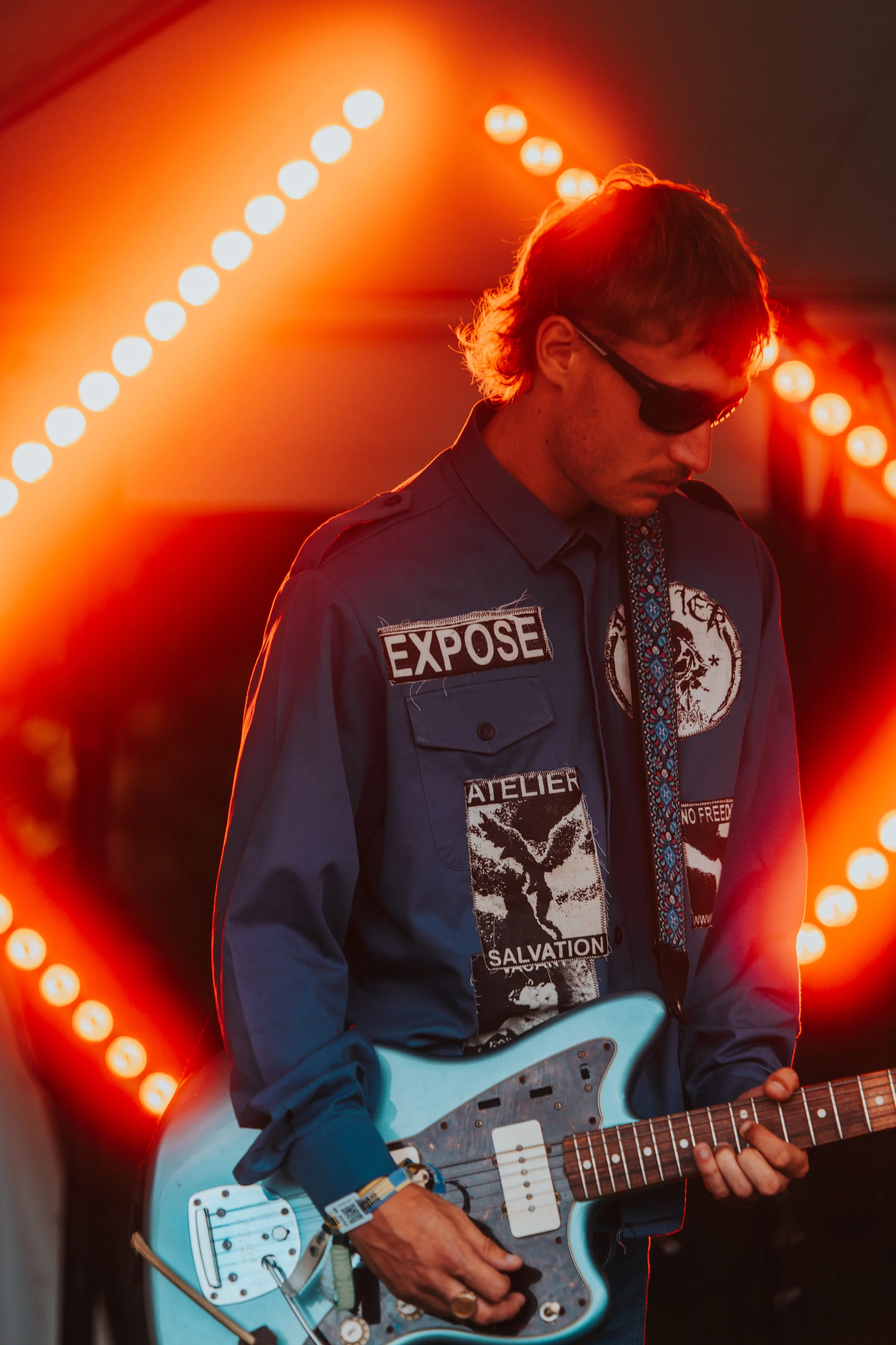 A man wearing sunglasses and a dark shirt with various patches, playing a light blue electric guitar, with orange stage lighting and circular lights in the background.