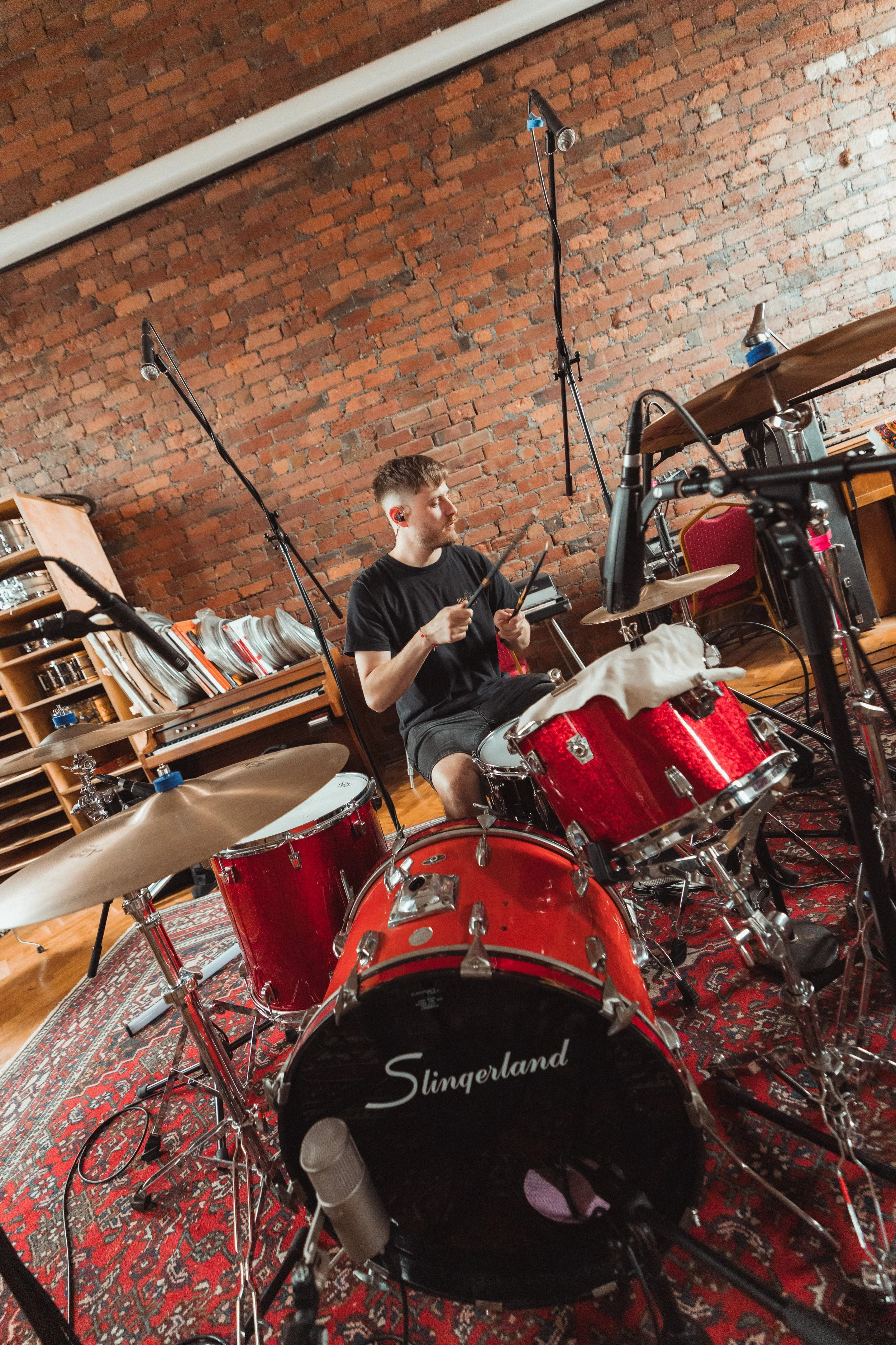A young man sitting behind a red drum set, holding drumsticks, in a room with an exposed brick wall, musical equipment, and a patterned rug.