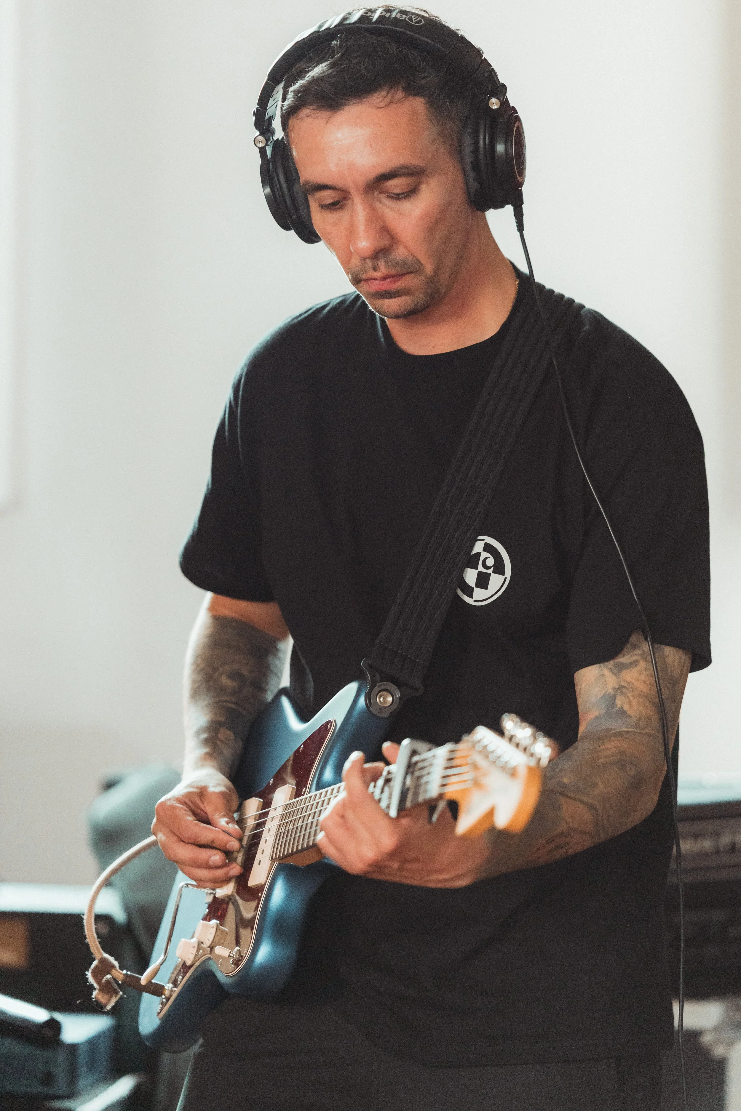 A man with tattoos wearing a black t-shirt and large headphones playing an electric guitar.