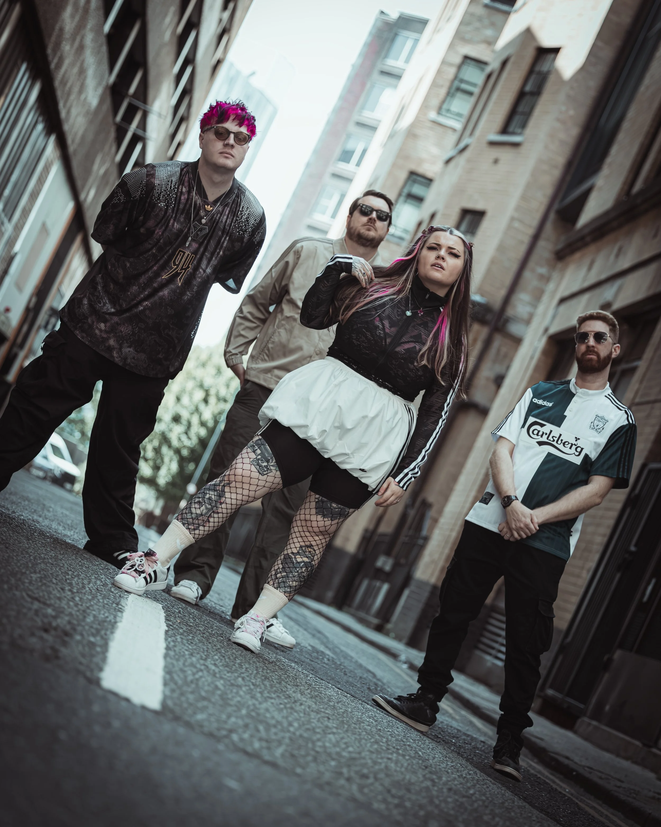 Four young adults standing and posing on a city street between tall buildings, wearing casual and stylish clothing, with one woman in the front playing with her hair and wearing fishnet tights.