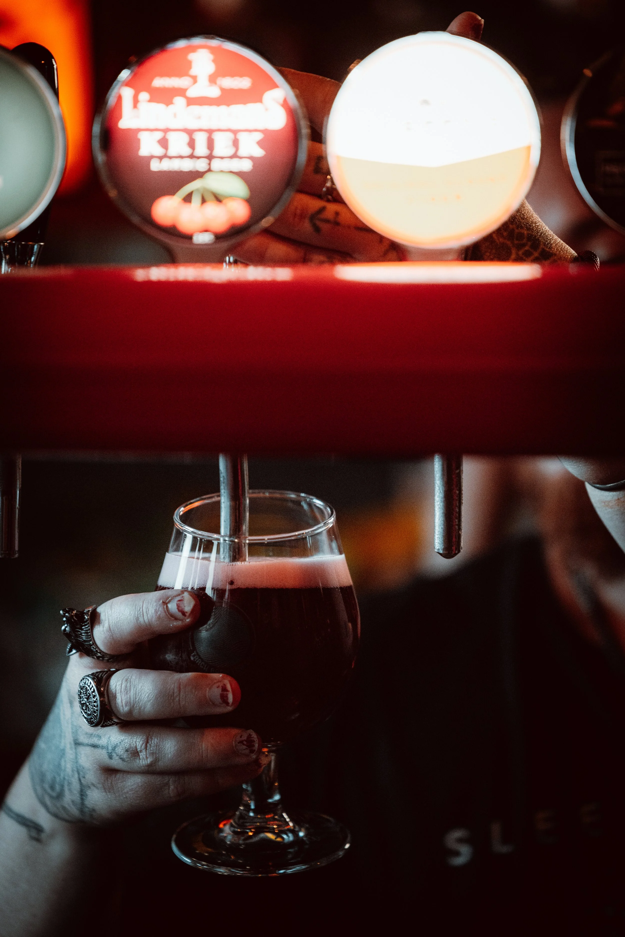 A person with tattoos on their hand is holding a glass of red wine. Above, there are shot glasses filled with different beverages, with one has a label that reads 'Lindenmann's Kriek' and an illustration of cherries.