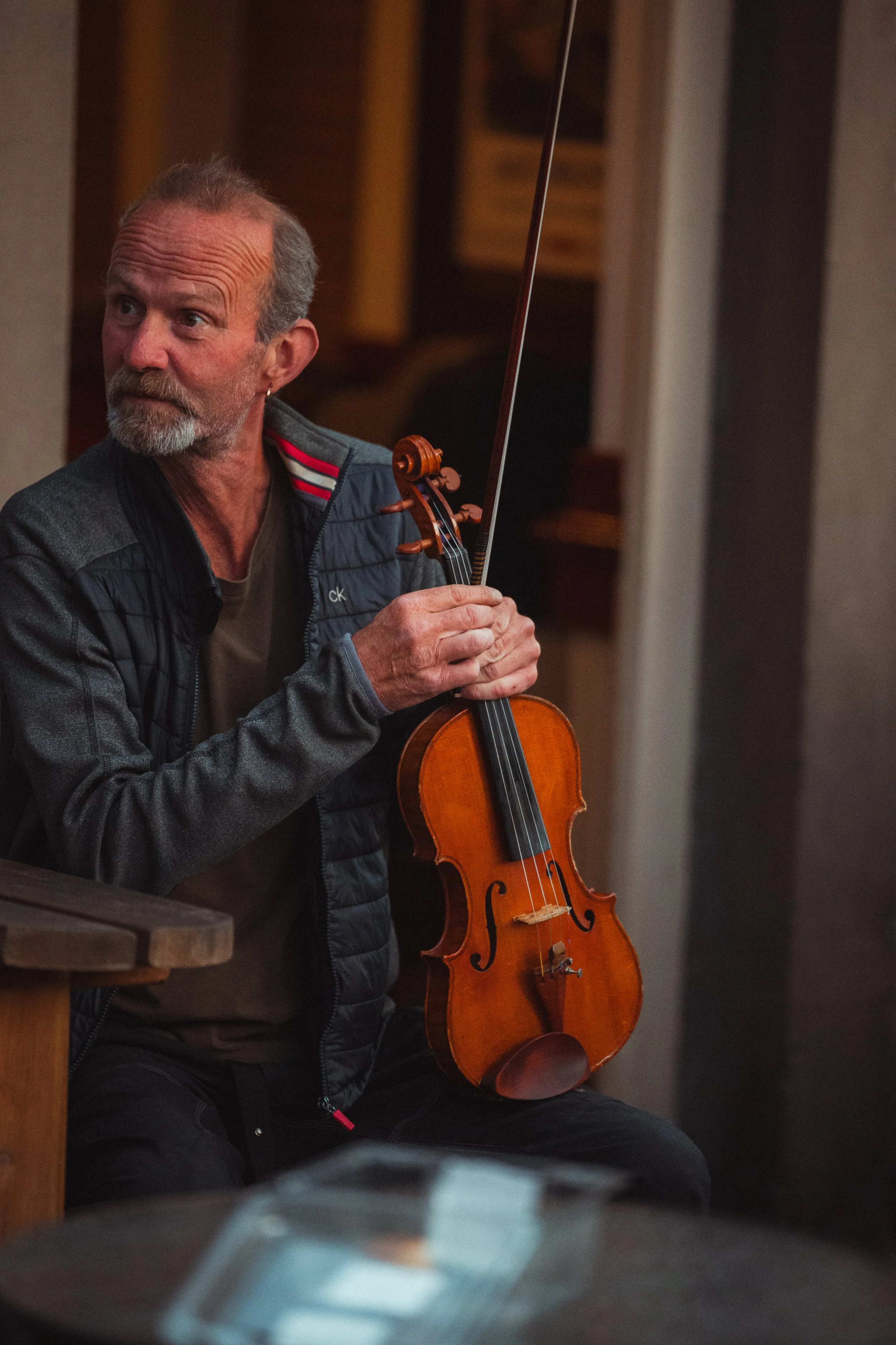 An older man with a beard and short gray hair holding a violin and bow, sitting in a room with wooden accents, wearing a gray jacket.