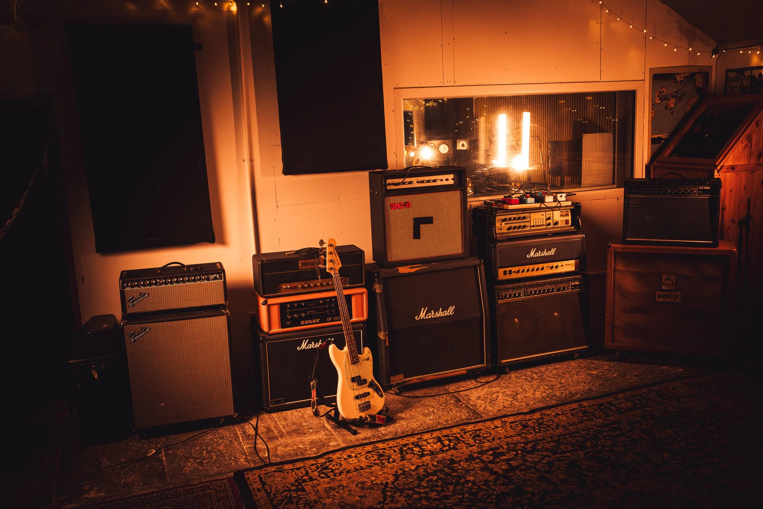 Music studio with various guitar amplifiers, a guitar, and audio equipment, illuminated by warm lighting and string lights, with a window overlooking another room.
