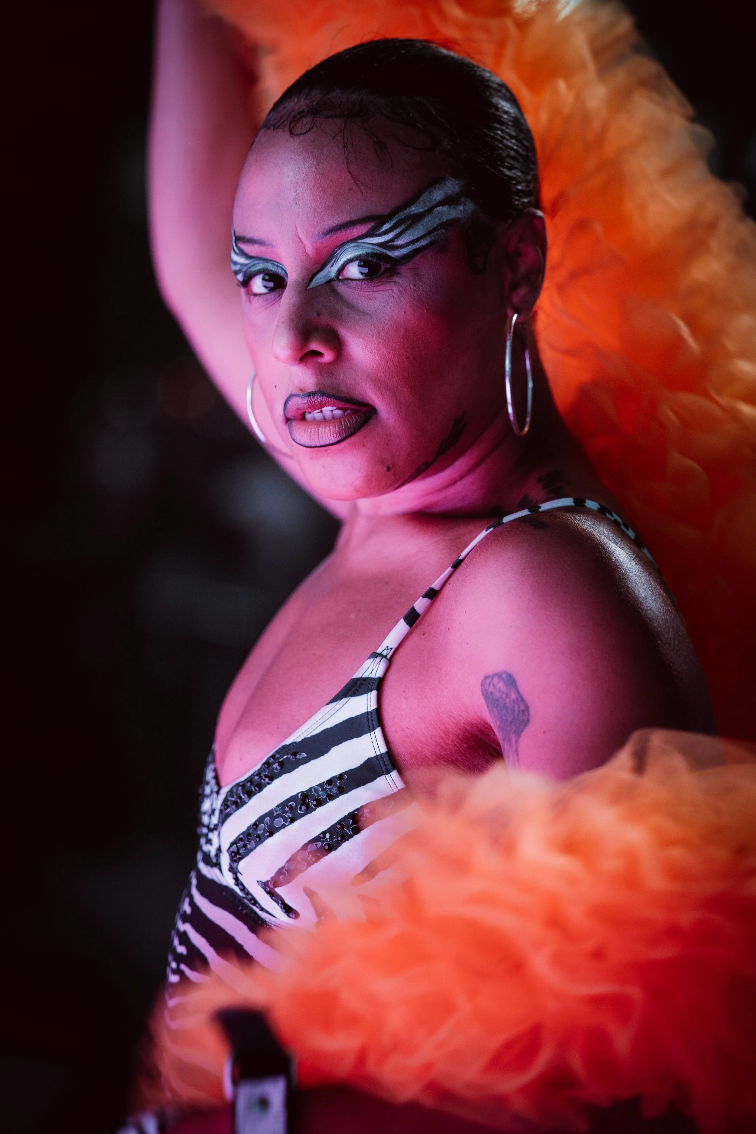 A woman with dark hair slicked back, wearing makeup with artistic eye designs, hoop earrings, and a striped top. She has a tattoo on her shoulder and a bright orange feathered accessory behind her. The background is dark.