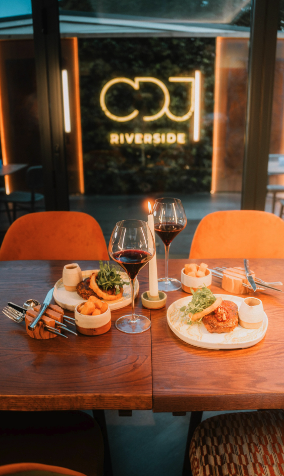 Elegant dining table set with two plates of food, two glasses of red wine, a lit candle, and orange chairs in front of a window with a neon sign reading 'RIVERSIDE.'