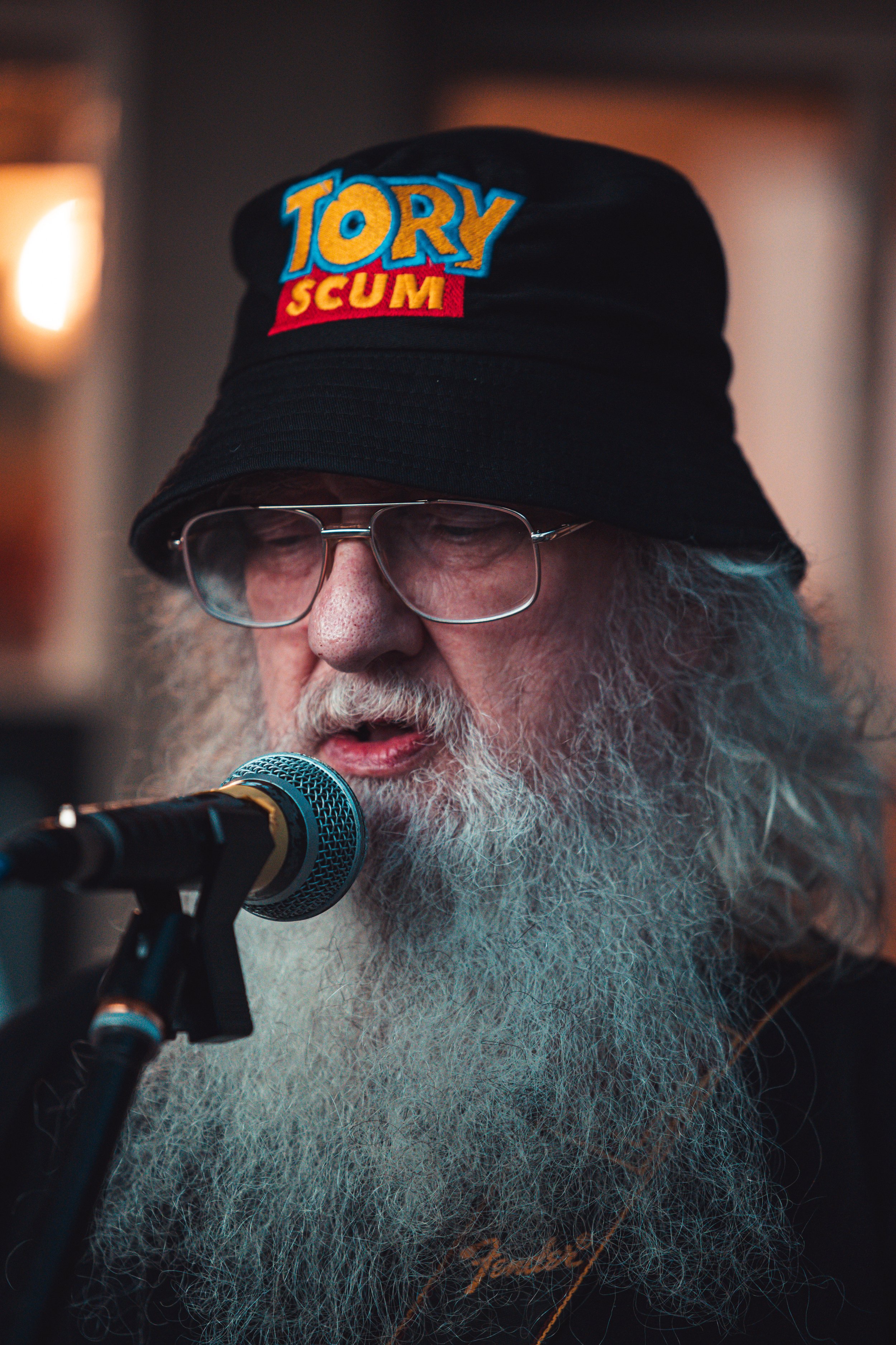 An elderly man with a long white beard and glasses, wearing a black bucket hat with colorful embroidery that reads 'TORY SCUM,' speaking into a microphone.