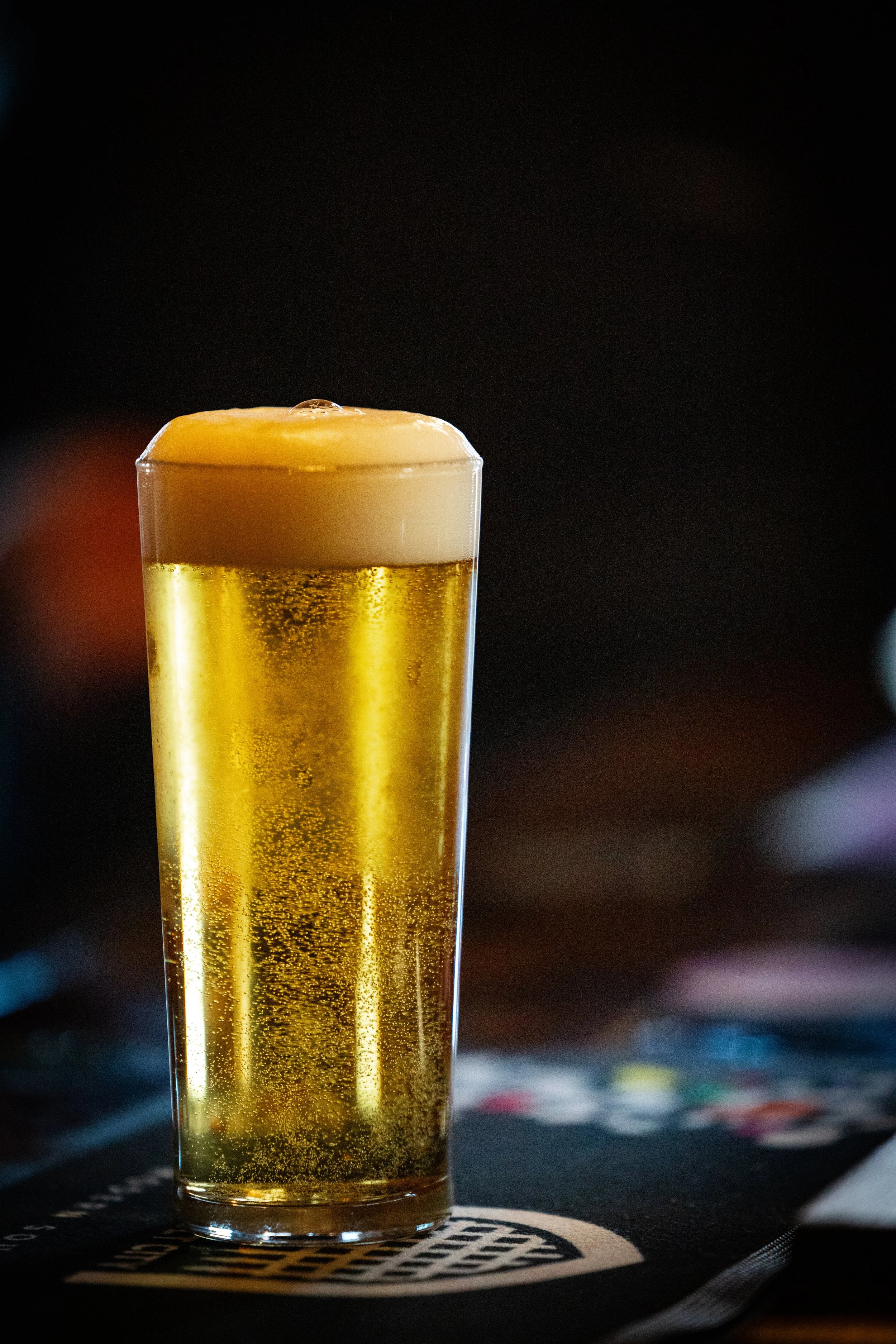 A tall glass of beer with foam on top, sitting on a dark bar or table surface.