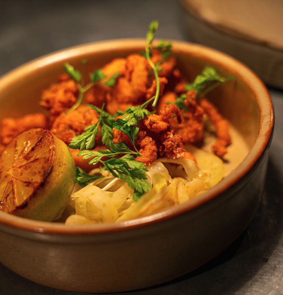 Fried chicken pieces garnished with fresh cilantro, served with a half grilled lime, on a beige bowl with shredded cabbage underneath.