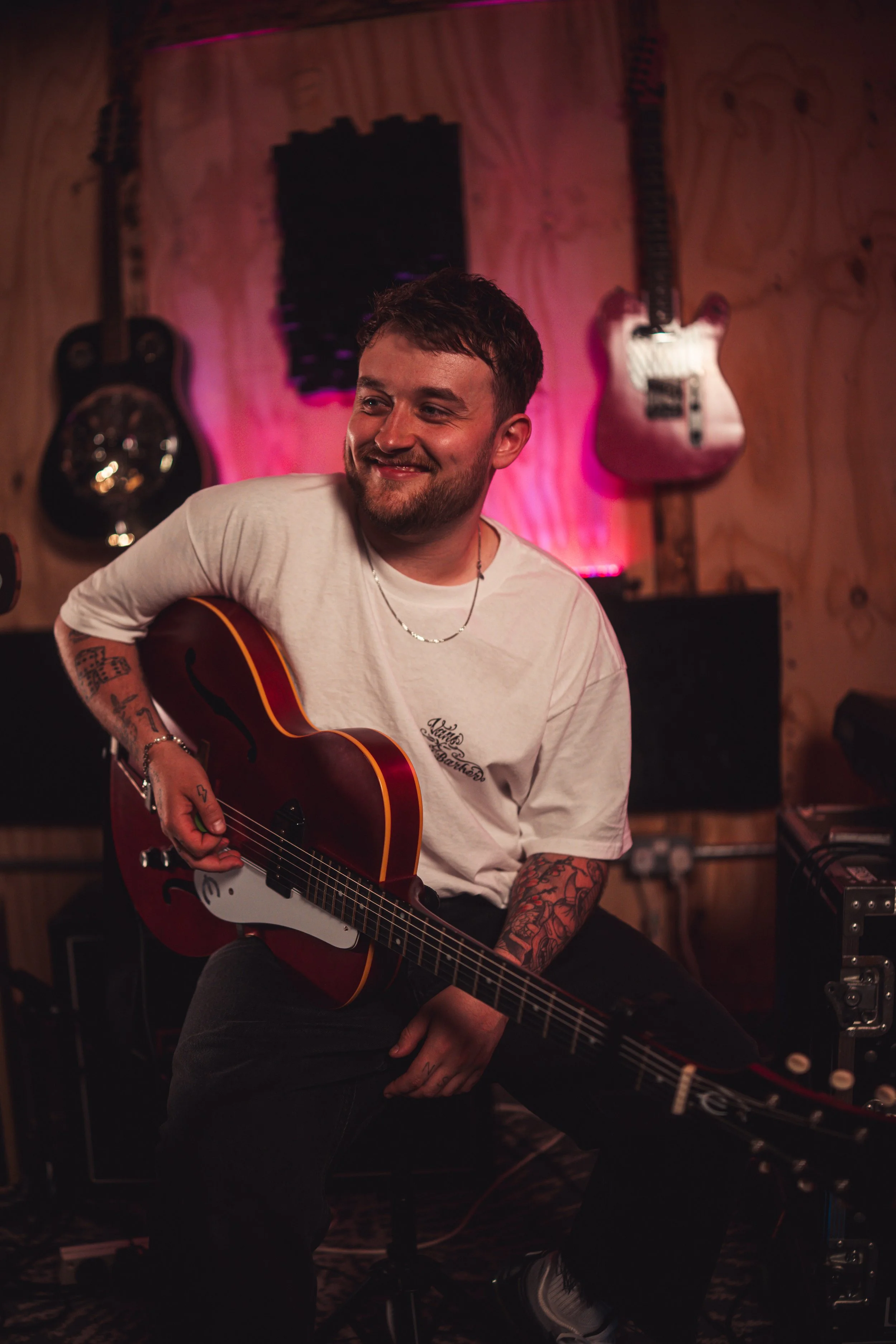 A young man with tattoos on his arms, smiling while playing an acoustic guitar in a music studio with wooden walls and guitars hanging in the background, illuminated by pink and purple lighting.