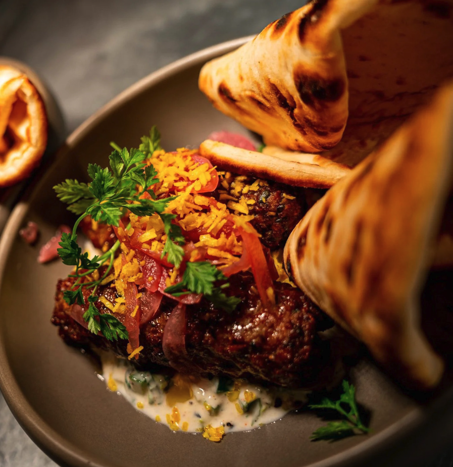 A gourmet dish featuring grilled flatbread, a seasoned beef patty topped with shredded cheese, cilantro, and pickled red onions, served with a creamy sauce, on a dark plate.