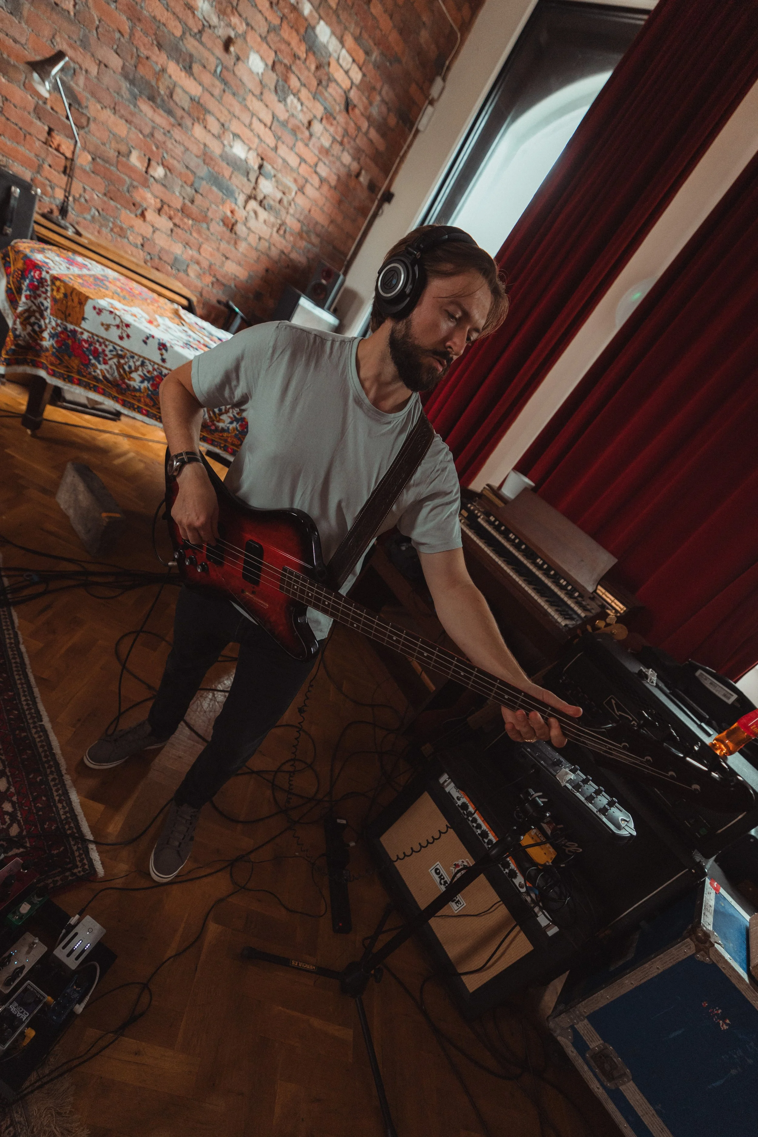A man with a beard and brown hair wears a gray t-shirt and black pants, playing an electric guitar in a music studio. He has black headphones on and is focused on playing. The studio has a brick wall, a window with red curtains, a patterned blanket o