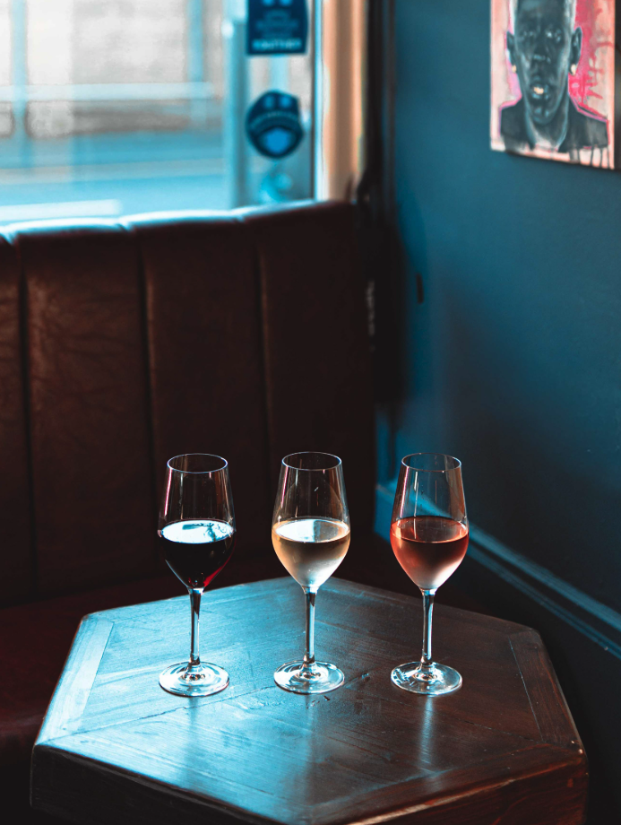 Three wine glasses with red, white, and rosé wine on a wooden table in a dimly lit bar or cafe, with a booth and a portrait on the wall in the background.