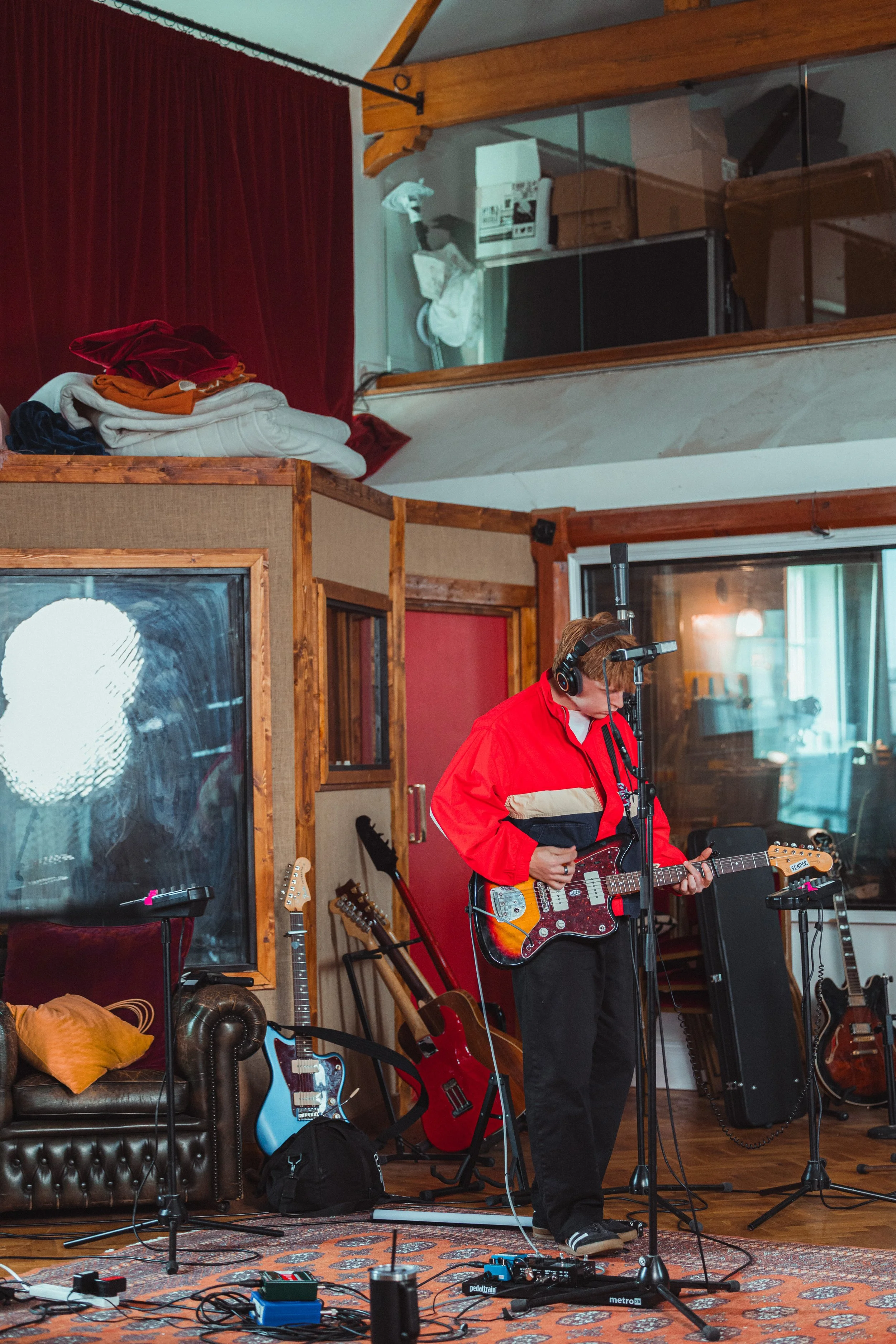 A person wearing a red and black jacket with headphones playing an electric guitar in a music studio. The studio has multiple guitars on stands, a sofa with a yellow pillow, and various music equipment and cables on the floor. There are wood and glas