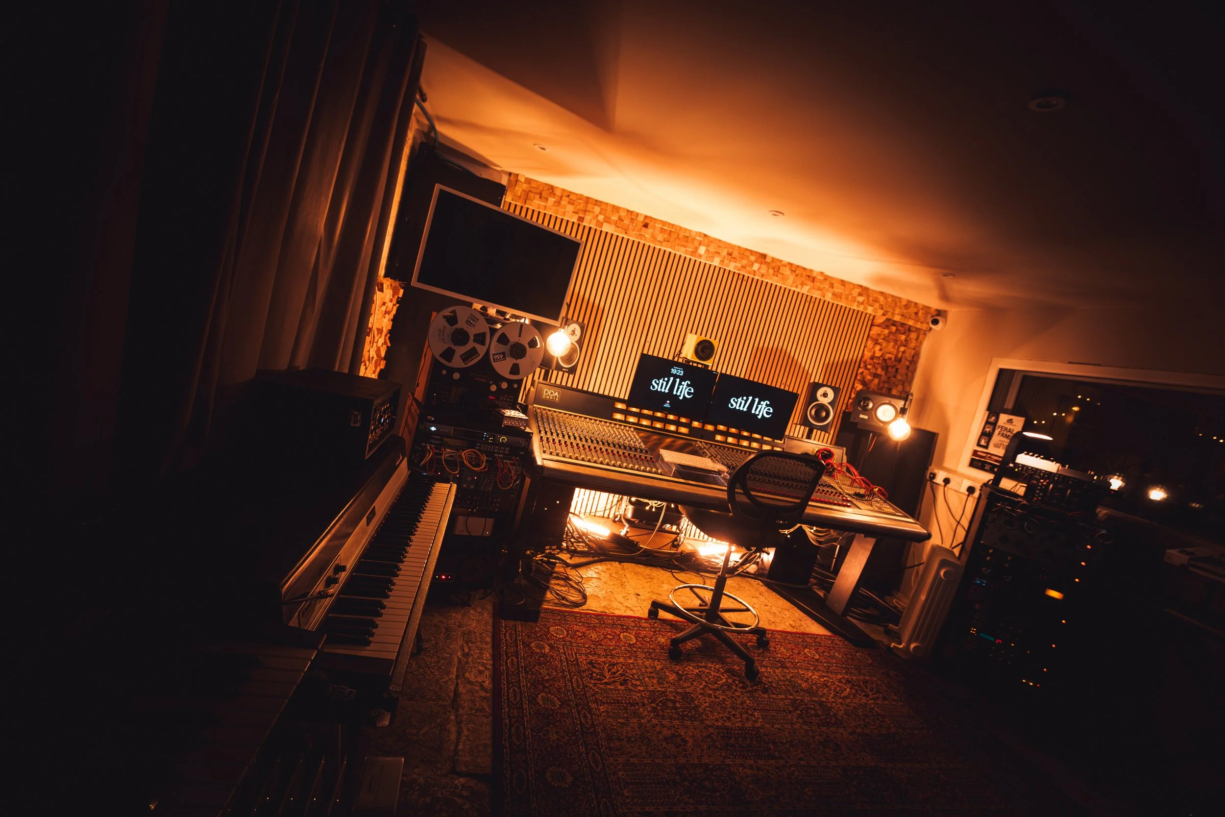 Recording studio with mixing console, computer screens displaying 'stil life', keyboard, speakers, reel-to-reel tape recorder, and musical equipment, illuminated by warm lighting.