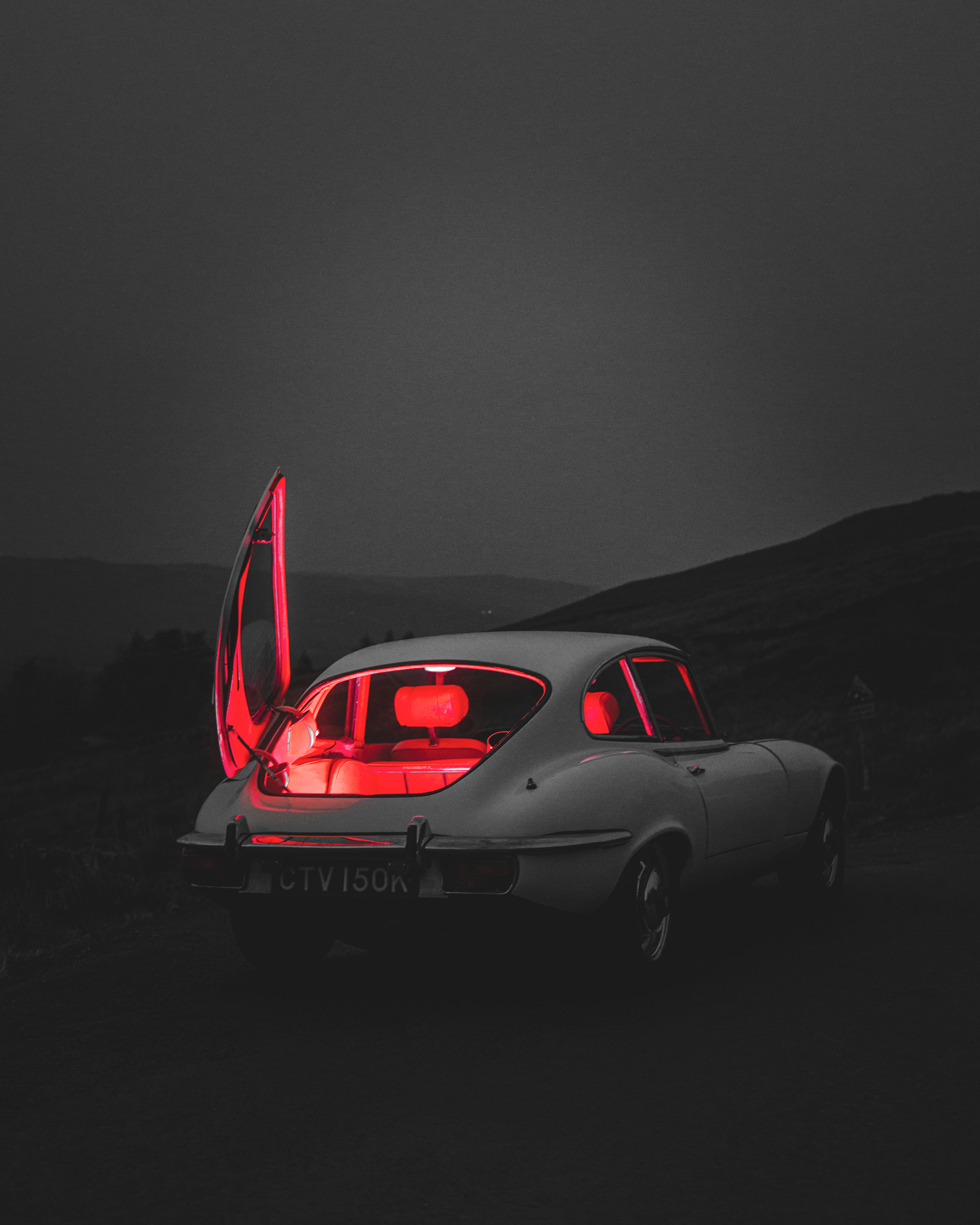 A vintage silver car with a neon red-lit interior and open gull-wing doors parked in a remote, dark landscape at night, with mountains in the background.