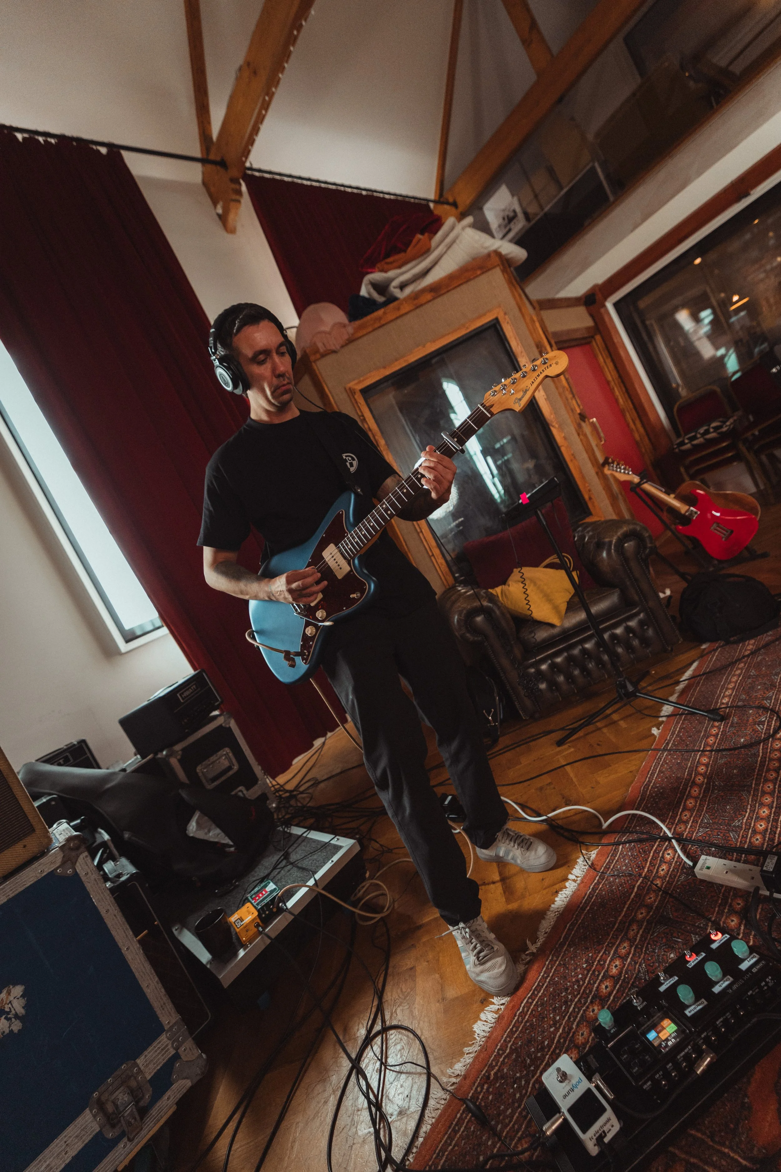 Man playing electric guitar in a recording studio, wearing headphones, with musical equipment and pedals around on the floor.