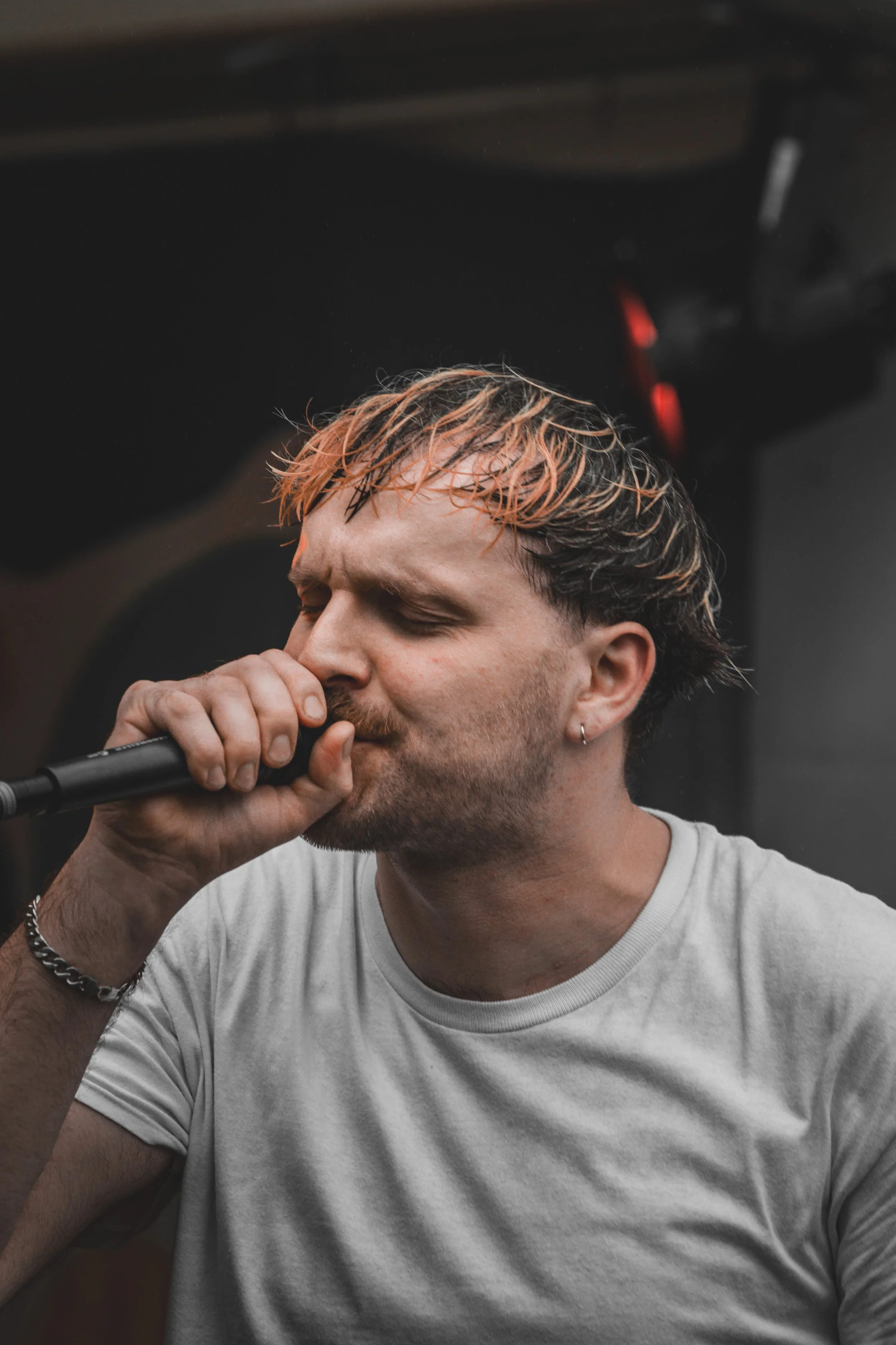 Man with light brown and blonde hair singing into a microphone, wearing a white T-shirt, with black background.