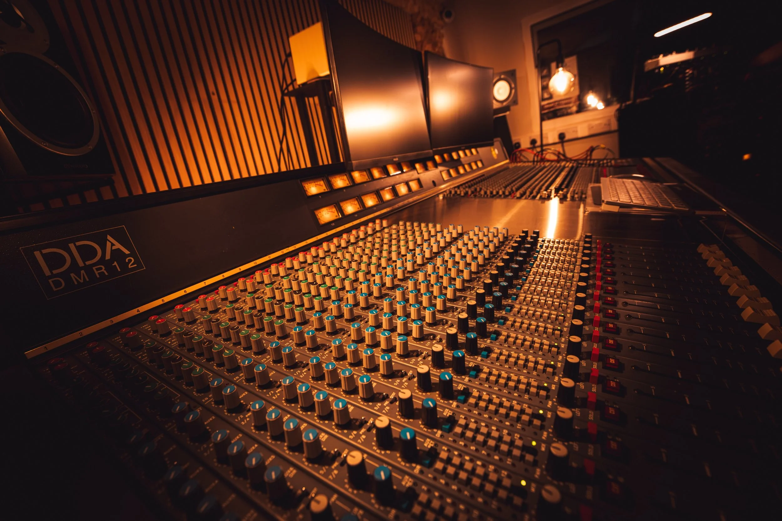 Recording studio room with a mixing console, multiple monitors, and studio speakers in a dimly lit environment with warm lighting.