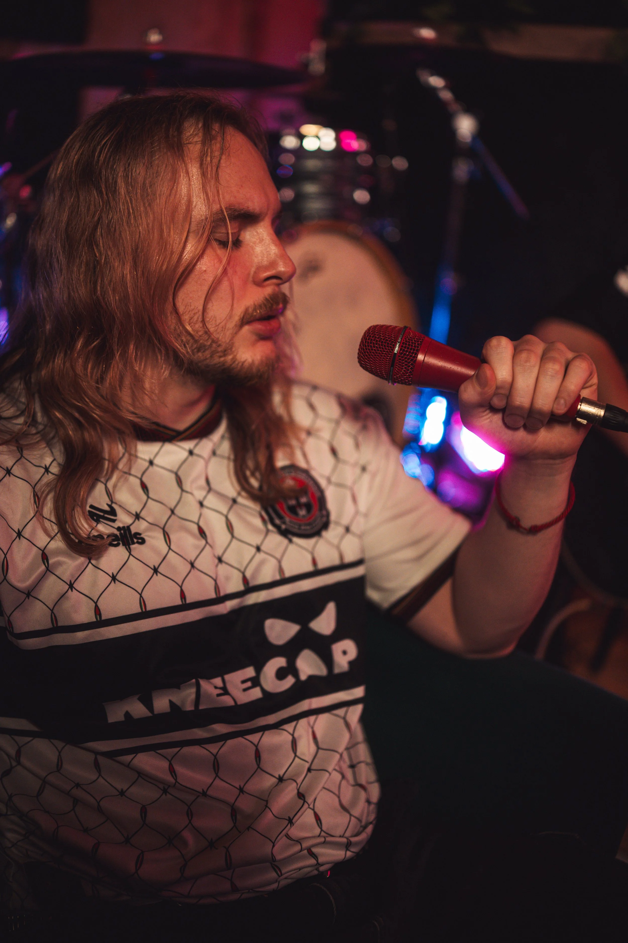 A man with long, wavy hair and a beard, singing into a red microphone, wearing a white shirt with a black geometric pattern and a logo, in a dimly lit setting with musical instruments in the background.