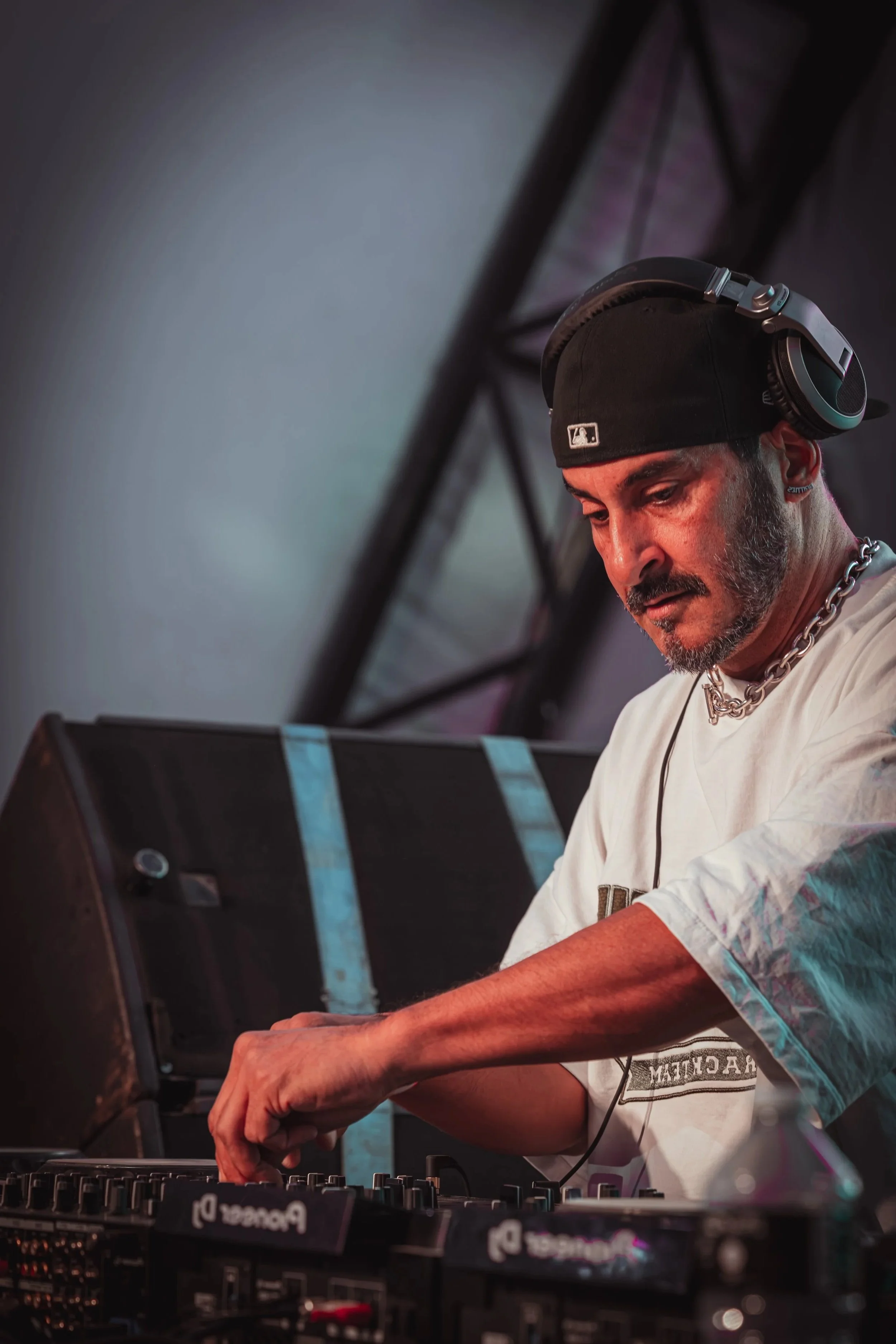 A DJ wearing a black baseball cap, a white t-shirt, and a chain necklace is working at a DJ booth, adjusting controls on a Pioneer DJ equipment with headphones on his head.