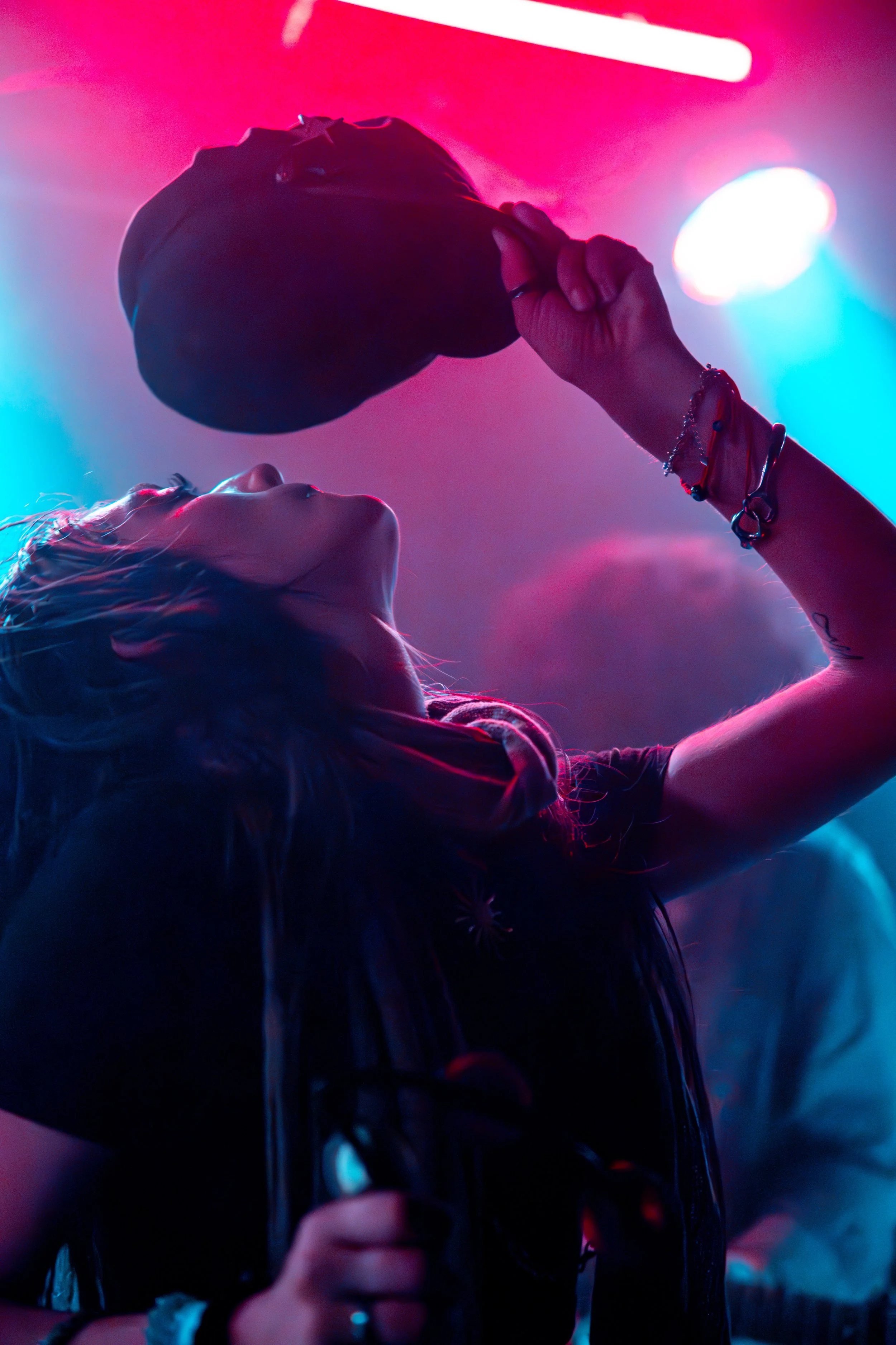 A person with long hair is holding a microphone close to their mouth at a concert or event, with pink and blue stage lighting creating a vibrant background.