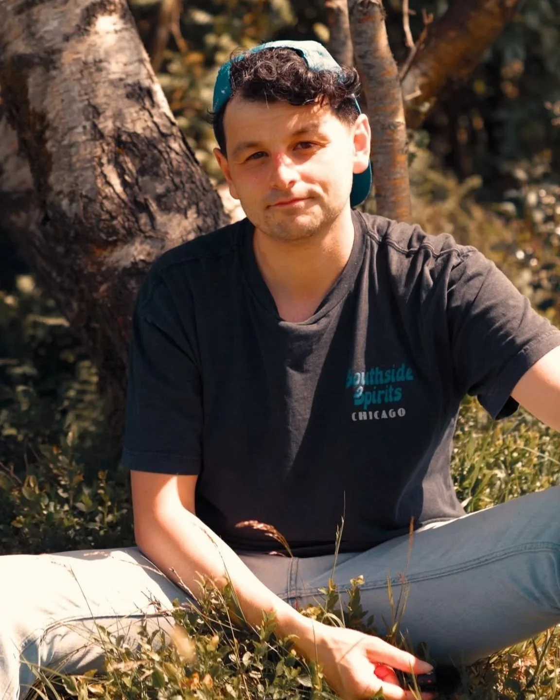 A young man with dark, curly hair wearing a black T-shirt and a backwards light blue cap sitting outdoors near a tree, surrounded by grass and foliage.