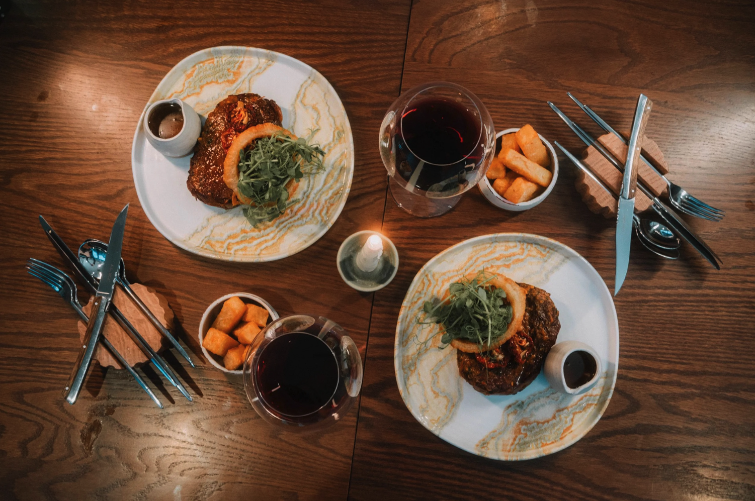 Two plates of gourmet burger with greens and onion rings, two glasses of red wine, side dishes of fried potato cubes, and cutlery on a wooden table with a candle in the center.