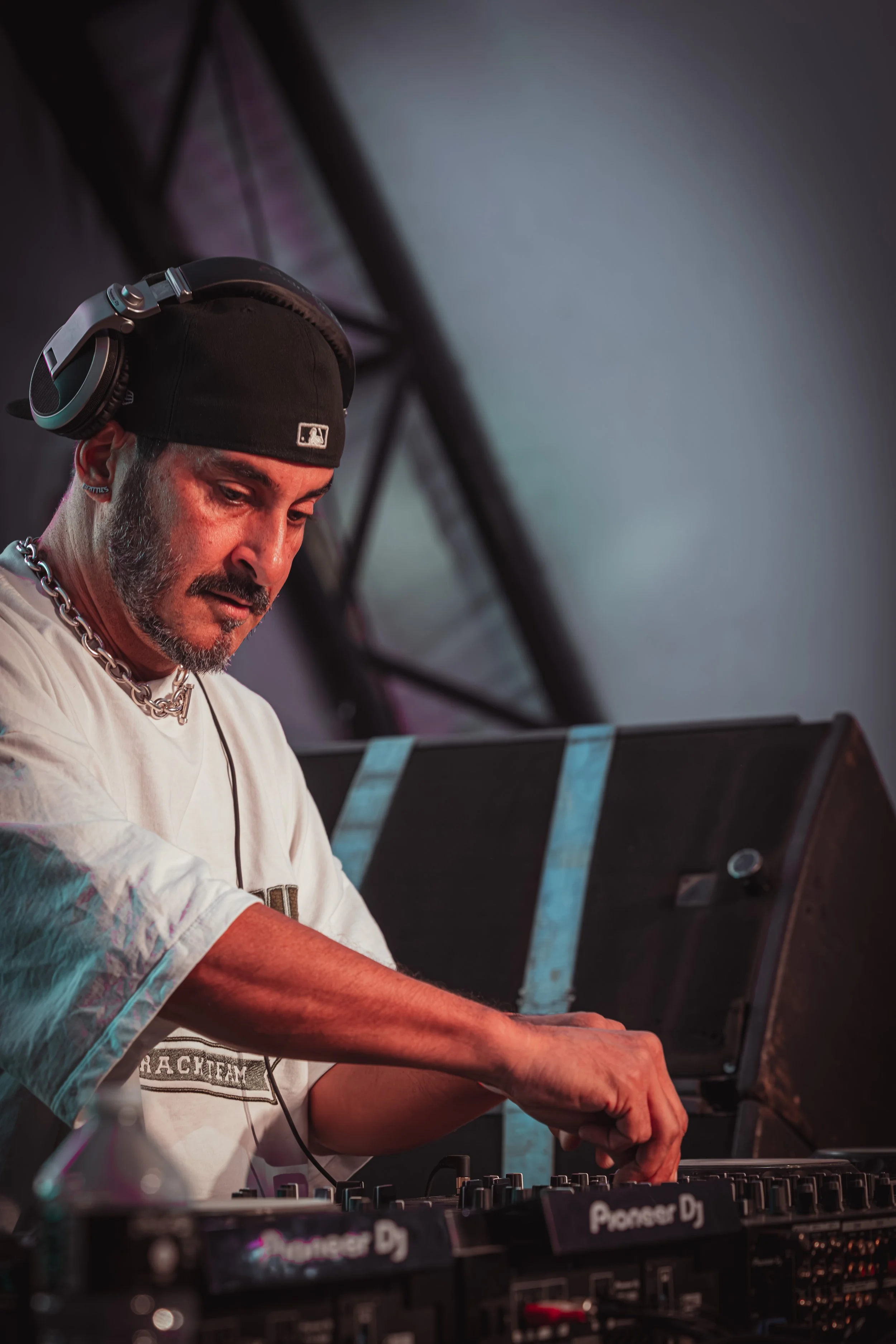 A DJ is performing at a music event, wearing a black cap, chain necklace, and white t-shirt, with headphones on, working with DJ equipment labeled 'Pioneer DJ'.