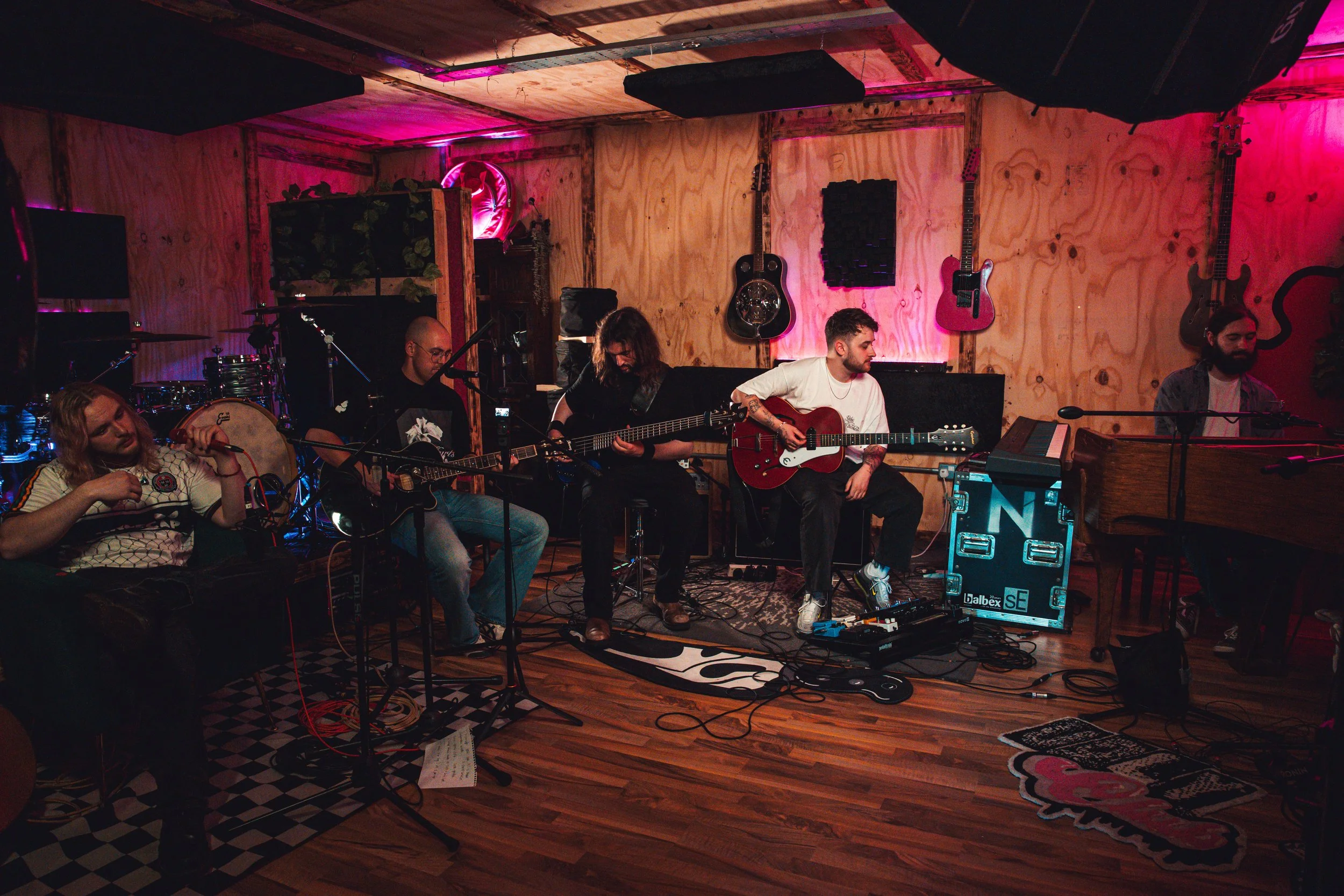 A band of five musicians performing in a cozy, dimly-lit room decorated with guitars on the wooden-paneled walls and pink lighting accents. They are playing guitars and drums while seated on chairs and a bench, surrounded by musical equipment.